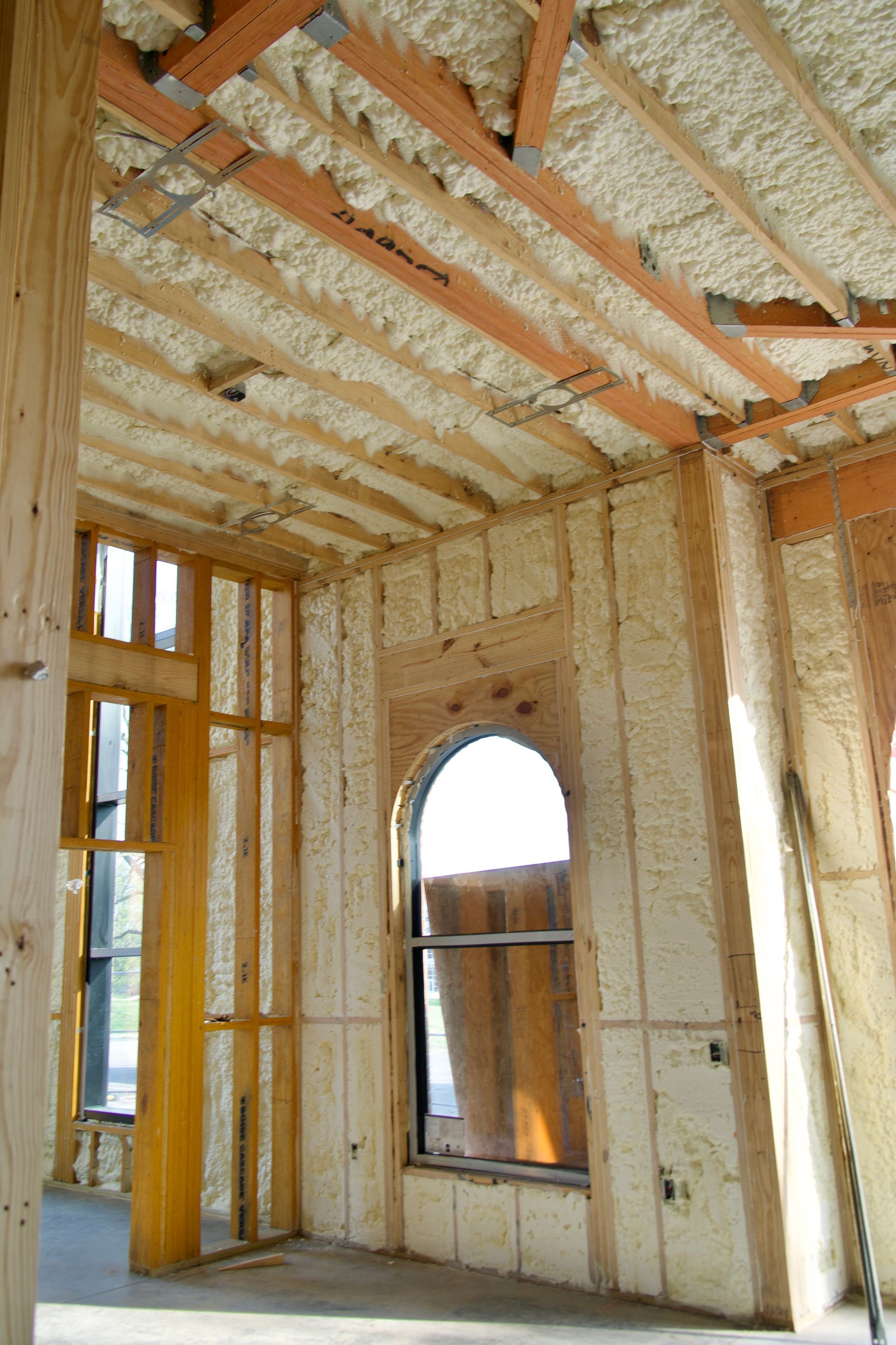 Interior view of a building frame with walls and ceiling joists insulated with spray foam, featuring an arched window.