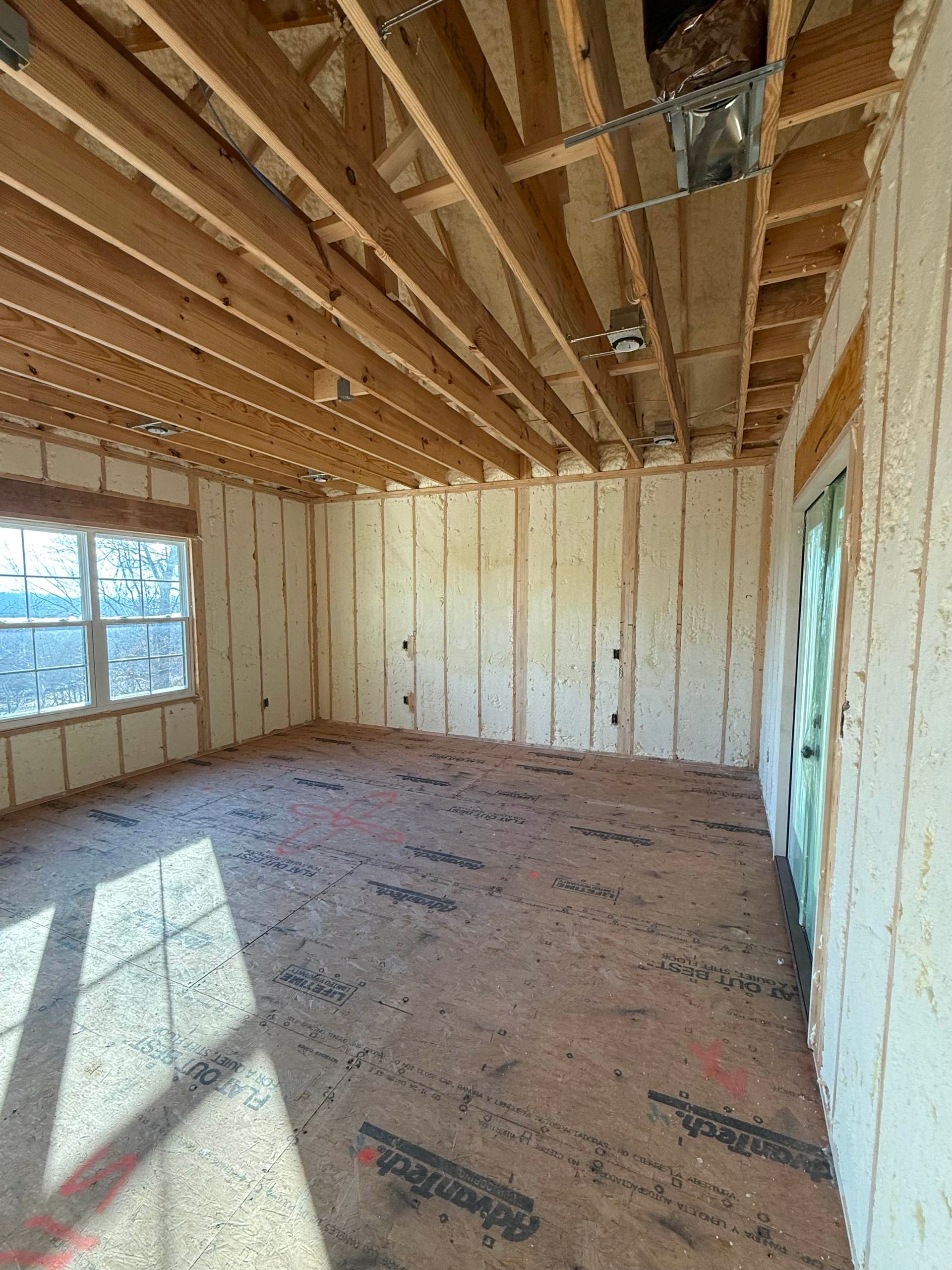 An unfinished room with exposed wooden rafters, light-colored spray foam insulation on the walls, and a window.