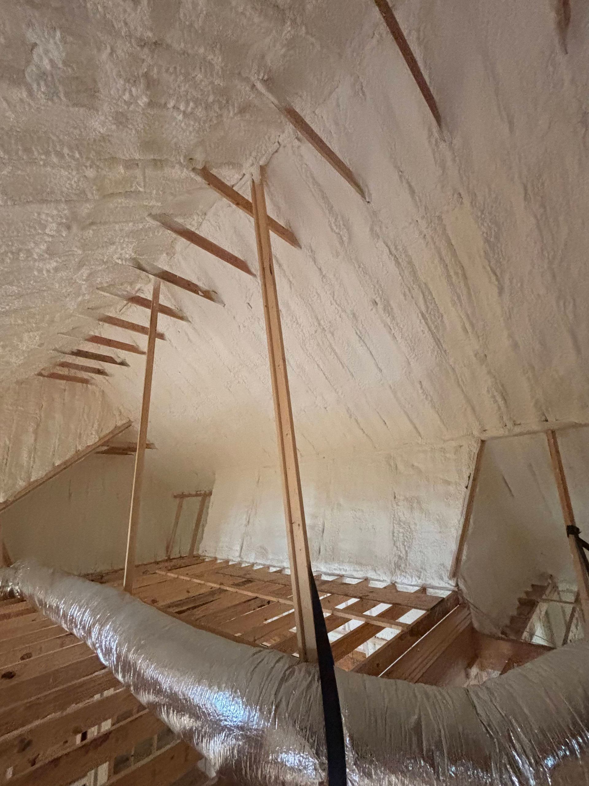 An attic space with spray foam insulation applied to the roof rafters and a metallic HVAC duct running across the floor.