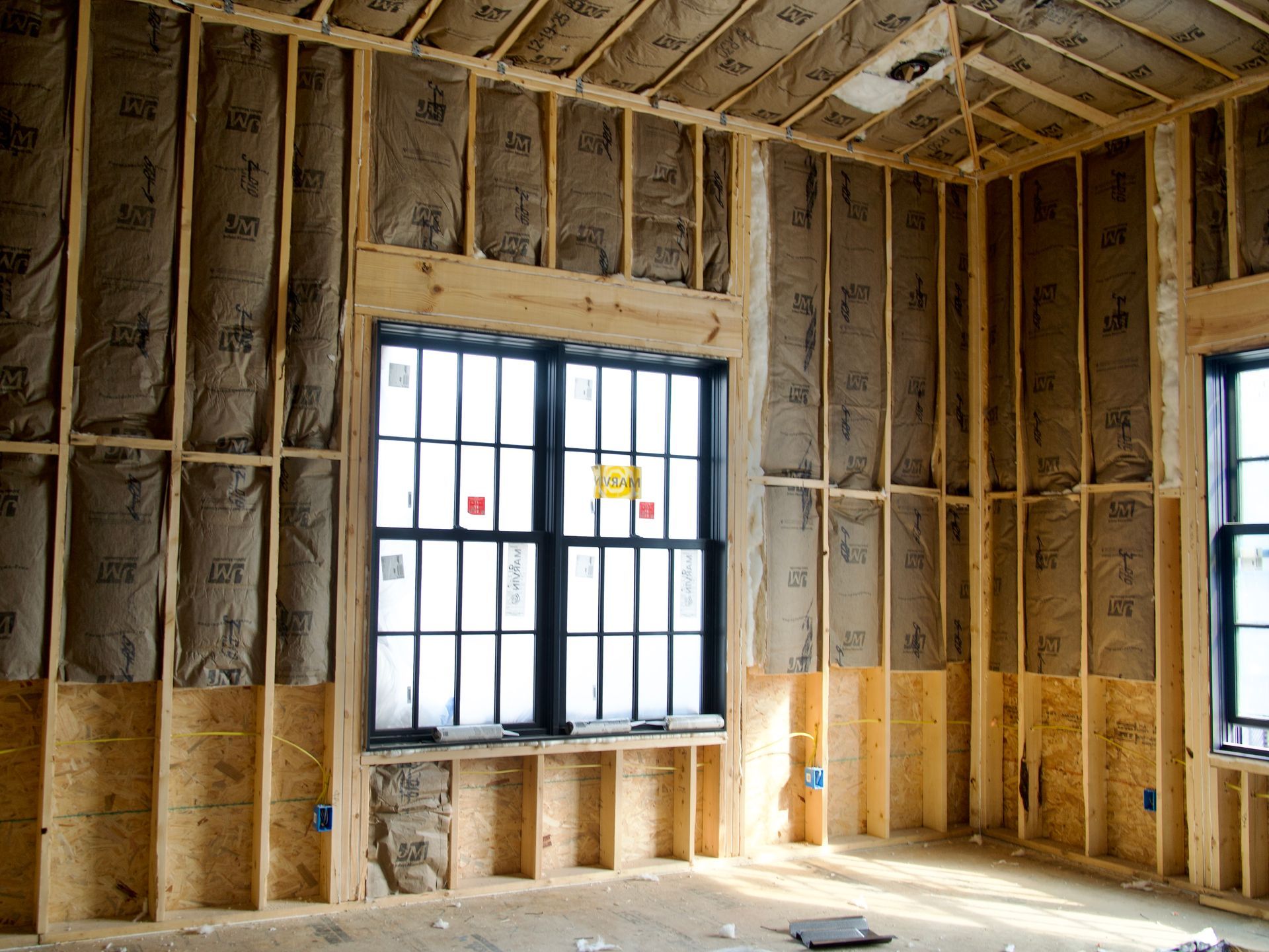 An interior view of a room under construction, featuring wooden wall studs, insulation batting, and two large windows.