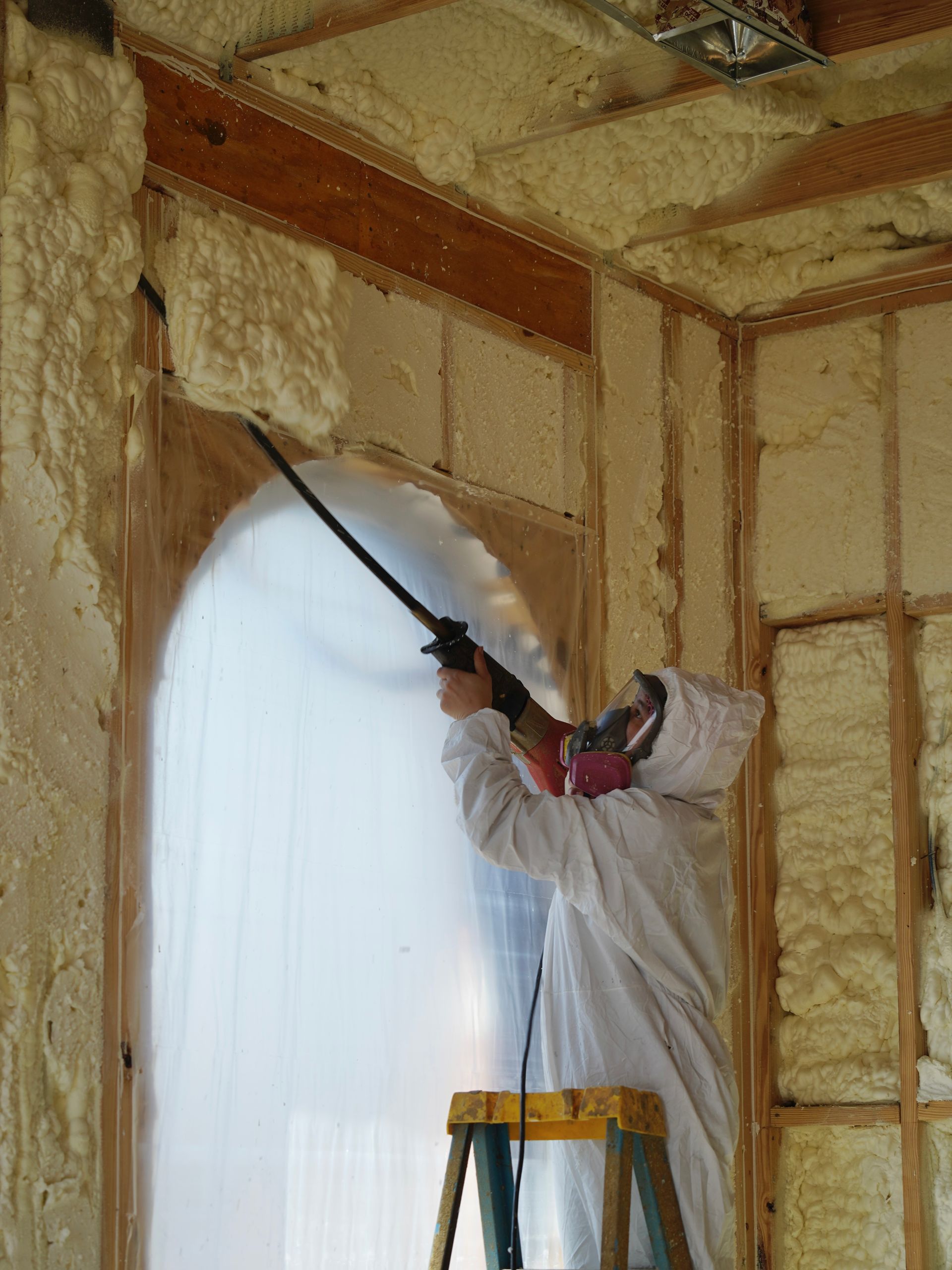 A worker in protective clothing stands on a ladder using a spray gun to apply yellow spray foam insulation to a wall.