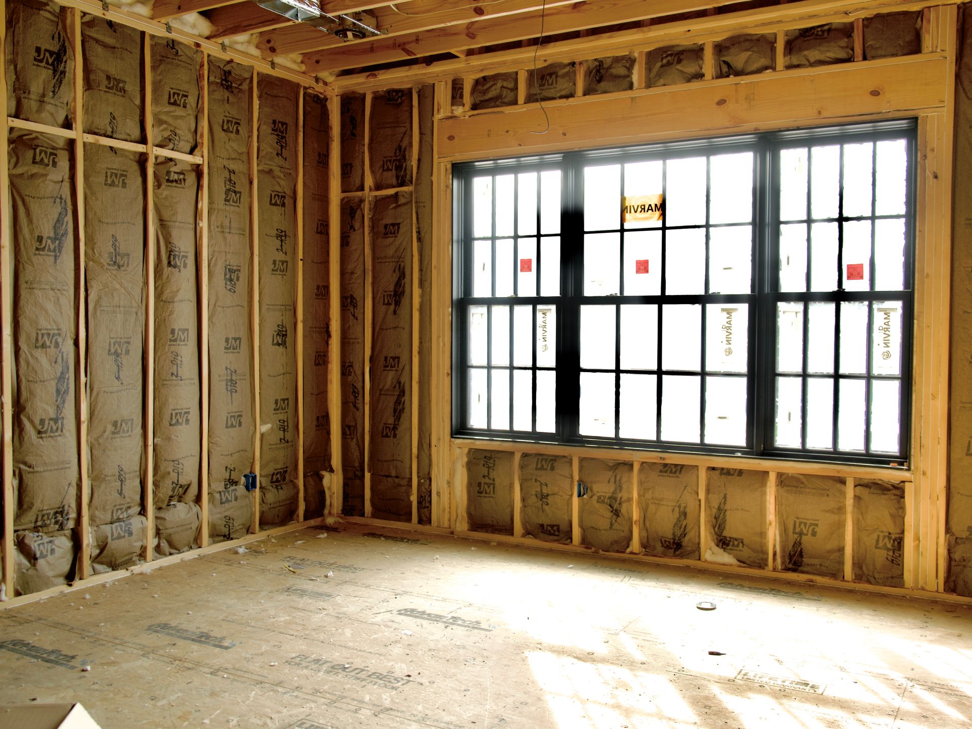 An interior room under construction with wood wall studs, installed insulation batts, and a large, black-framed window.