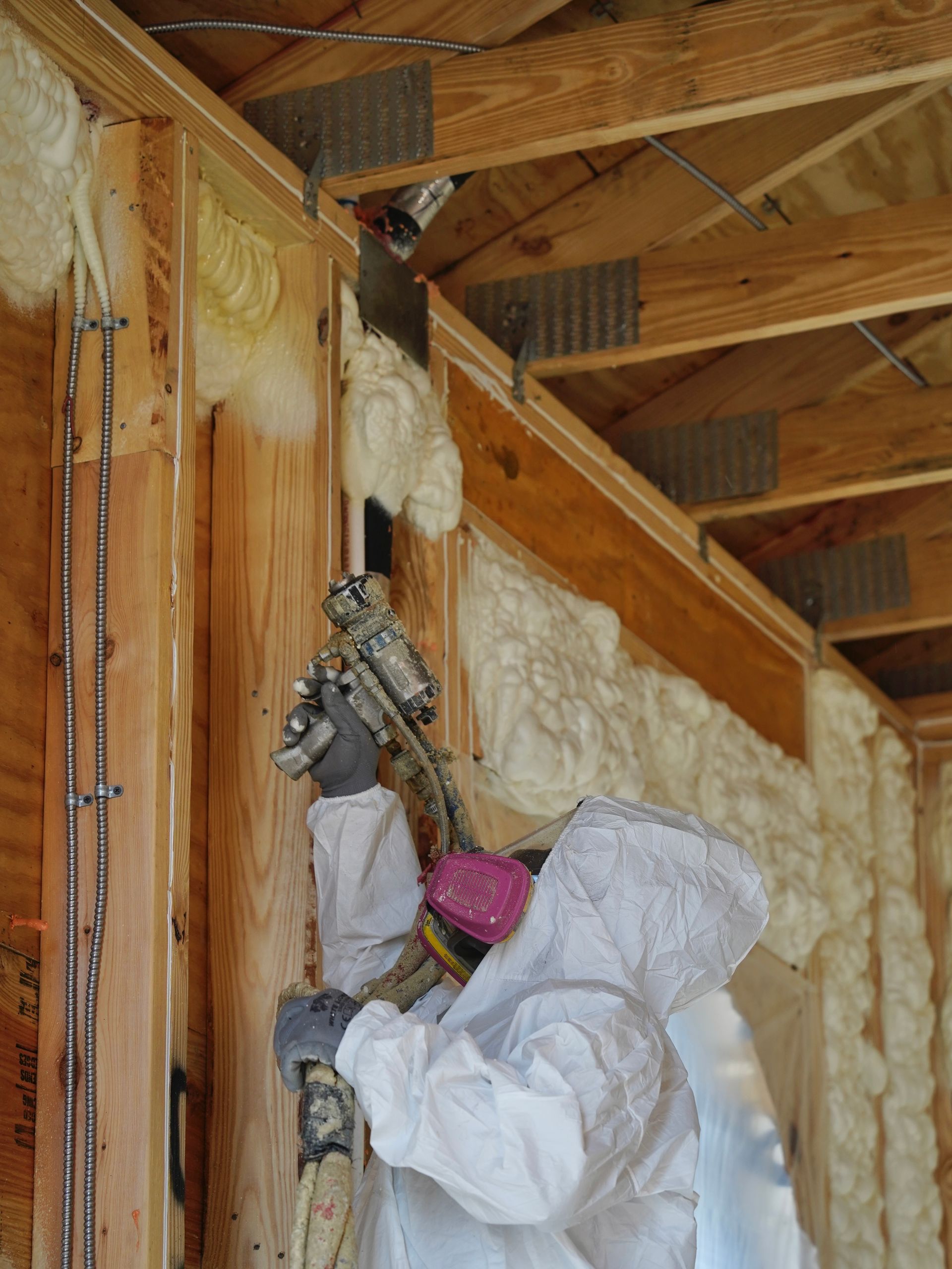 A person in protective coveralls and a respirator uses a spray gun to apply insulation foam to wooden wall studs.