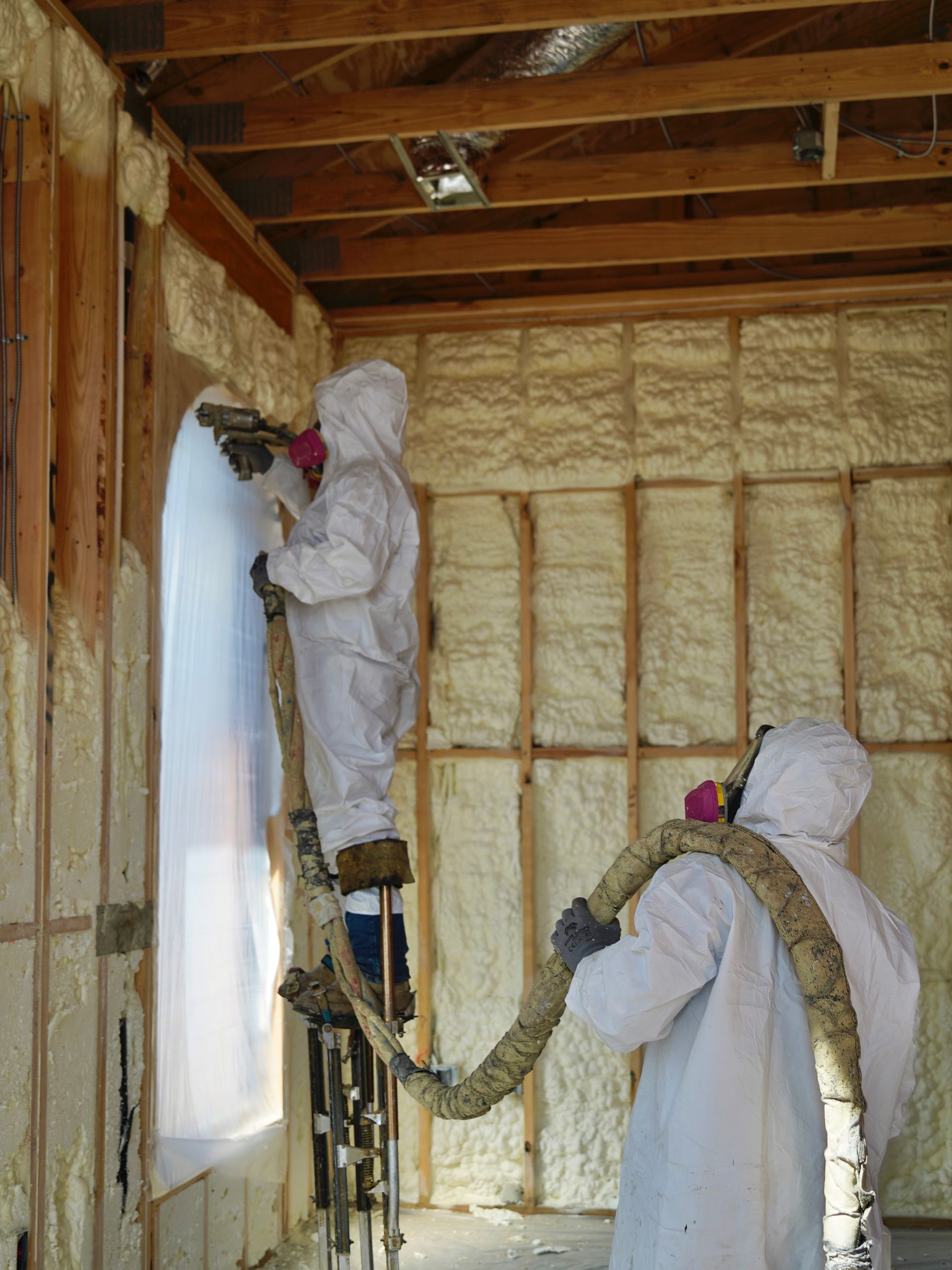 Two workers in protective suits and respirators spray insulation foam onto wooden wall studs inside a building.