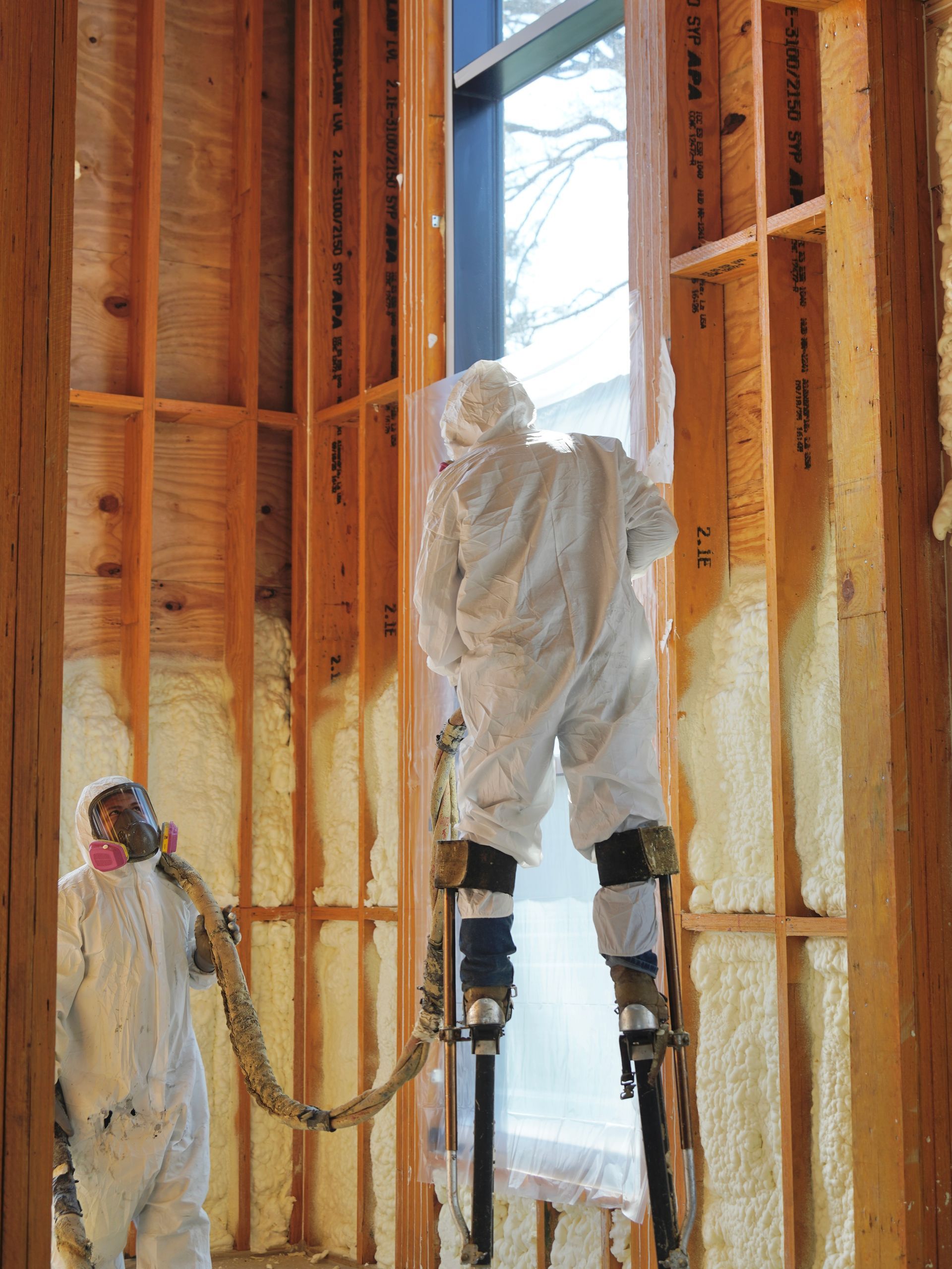 Two workers in protective suits apply spray foam insulation to wooden wall studs, with one using stilts to reach high areas.