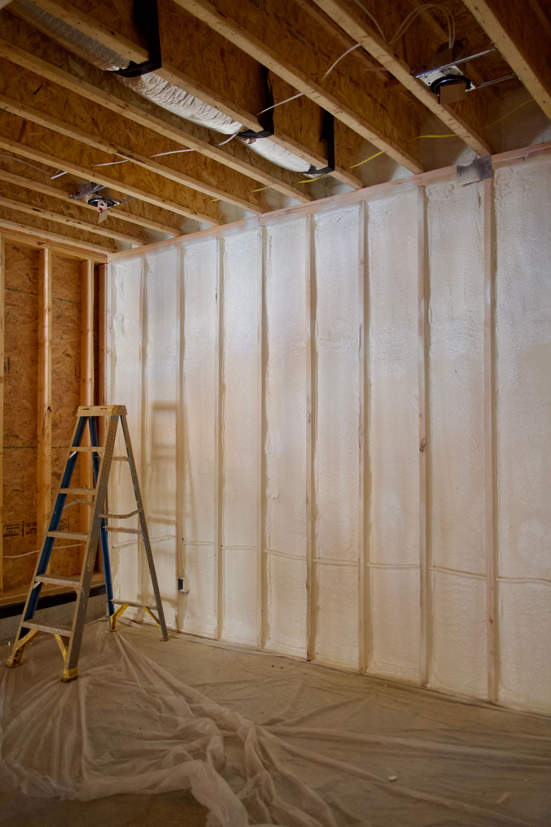 An unfinished room with a stud wall insulated with white spray foam, a ladder, and a plastic-covered floor.