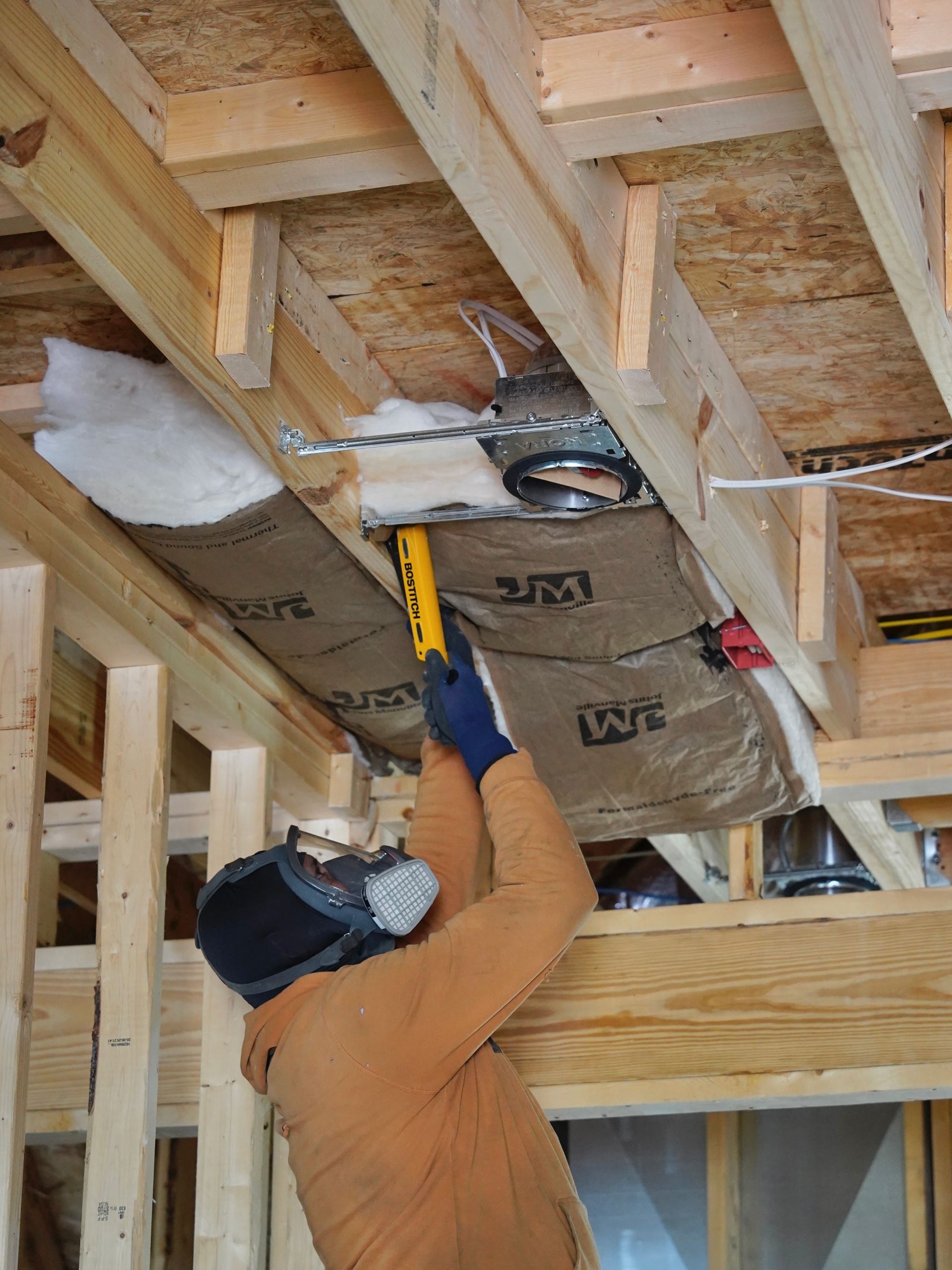 A worker in a respirator installs 3M fire barrier insulation around a ceiling light fixture in a wooden frame building.