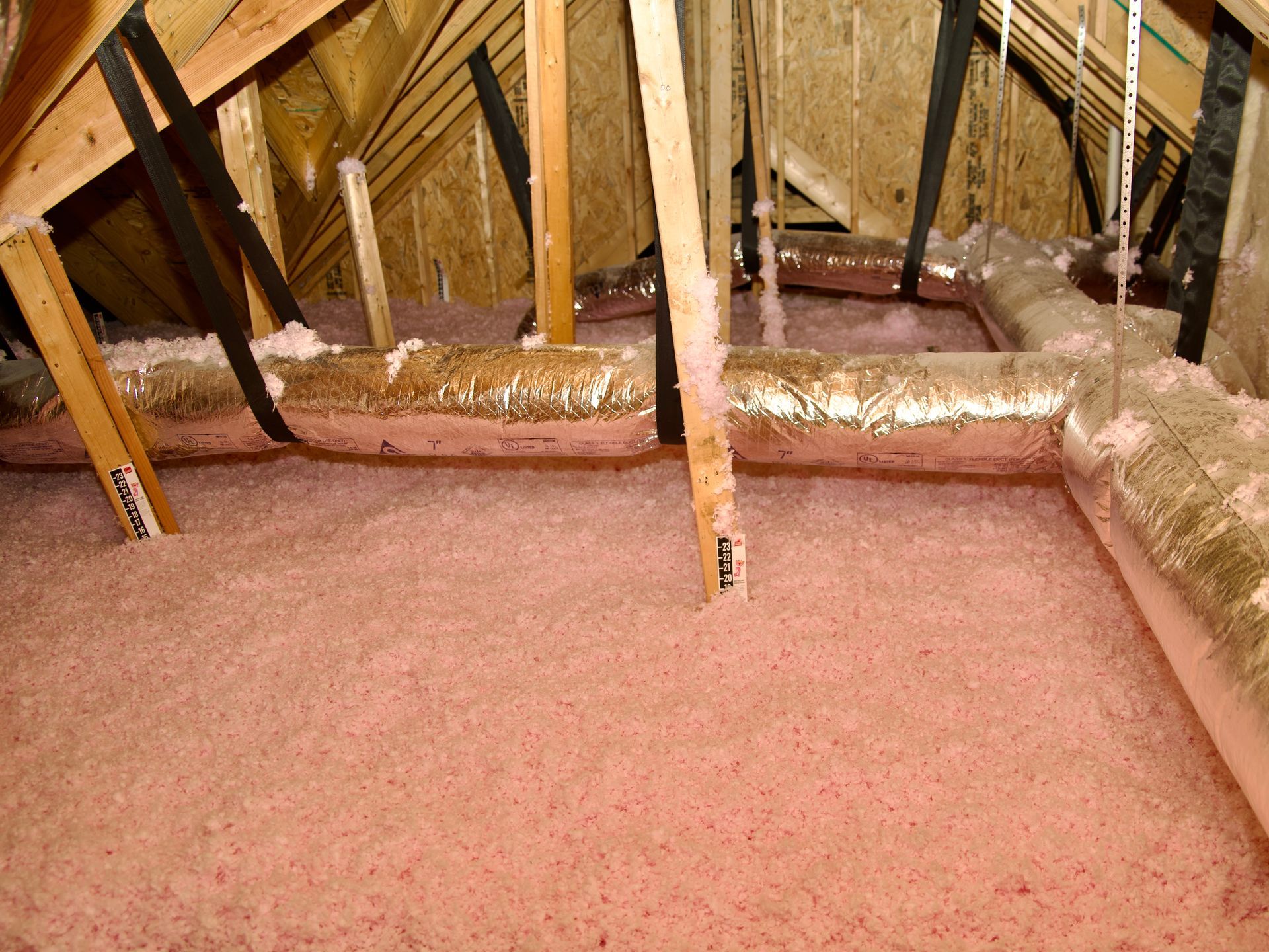 An attic space featuring pink loose-fill insulation between wooden rafters and insulated ventilation ducts.