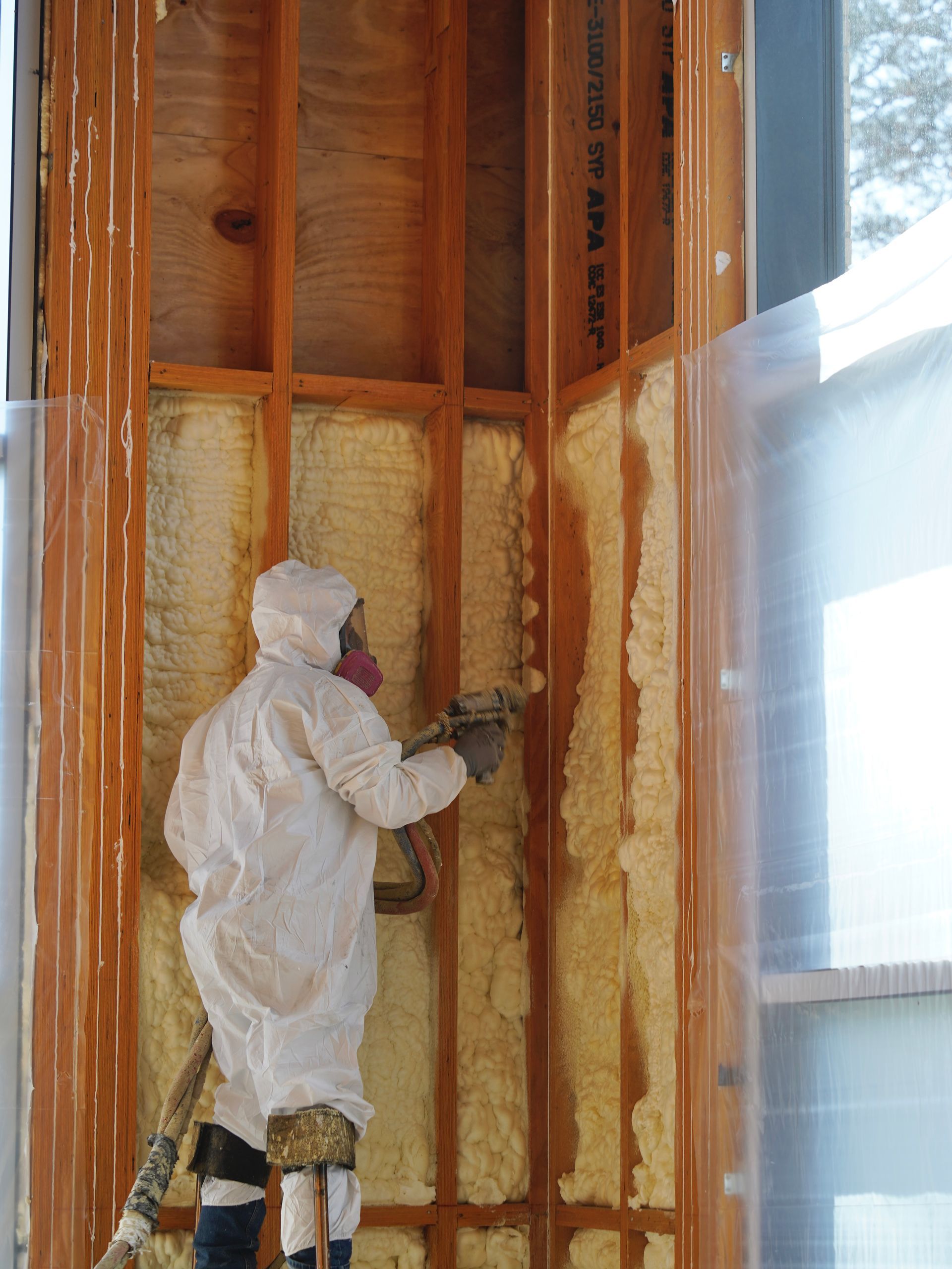A worker in full protective gear sprays yellow expanding foam insulation into wall studs at a construction site.