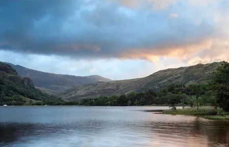 Lake surrounded by green hills under a cloudy sky with orange and blue hues.