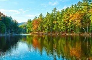 Lush green and autumn-hued trees line a calm lake, reflecting the foliage and blue sky.