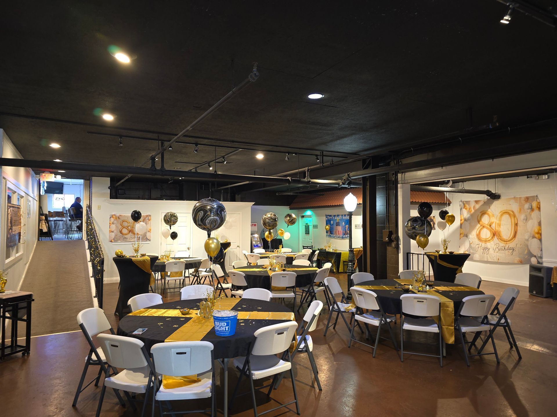 Event space decorated for a party with black and gold. Round tables with chairs, balloons, and artwork on walls.