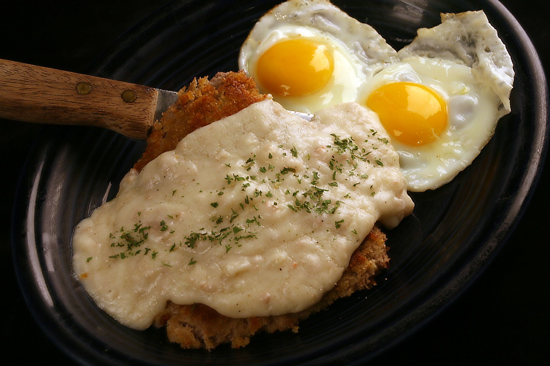 Fried cutlet with gravy and two sunny-side-up eggs on a black plate, wooden spatula on the side.