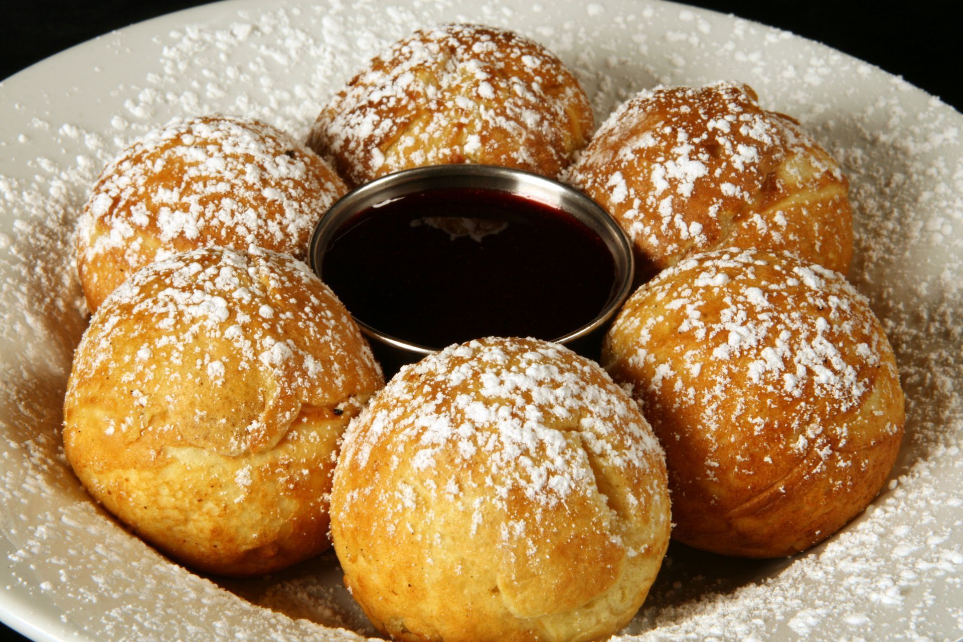 Six golden-brown beignets dusted with powdered sugar, arranged around a small dish of dark sauce on a white plate.