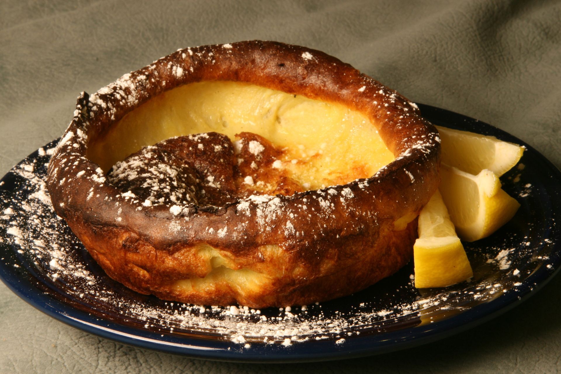 Dutch baby pancake with powdered sugar and lemon wedges on blue plate.