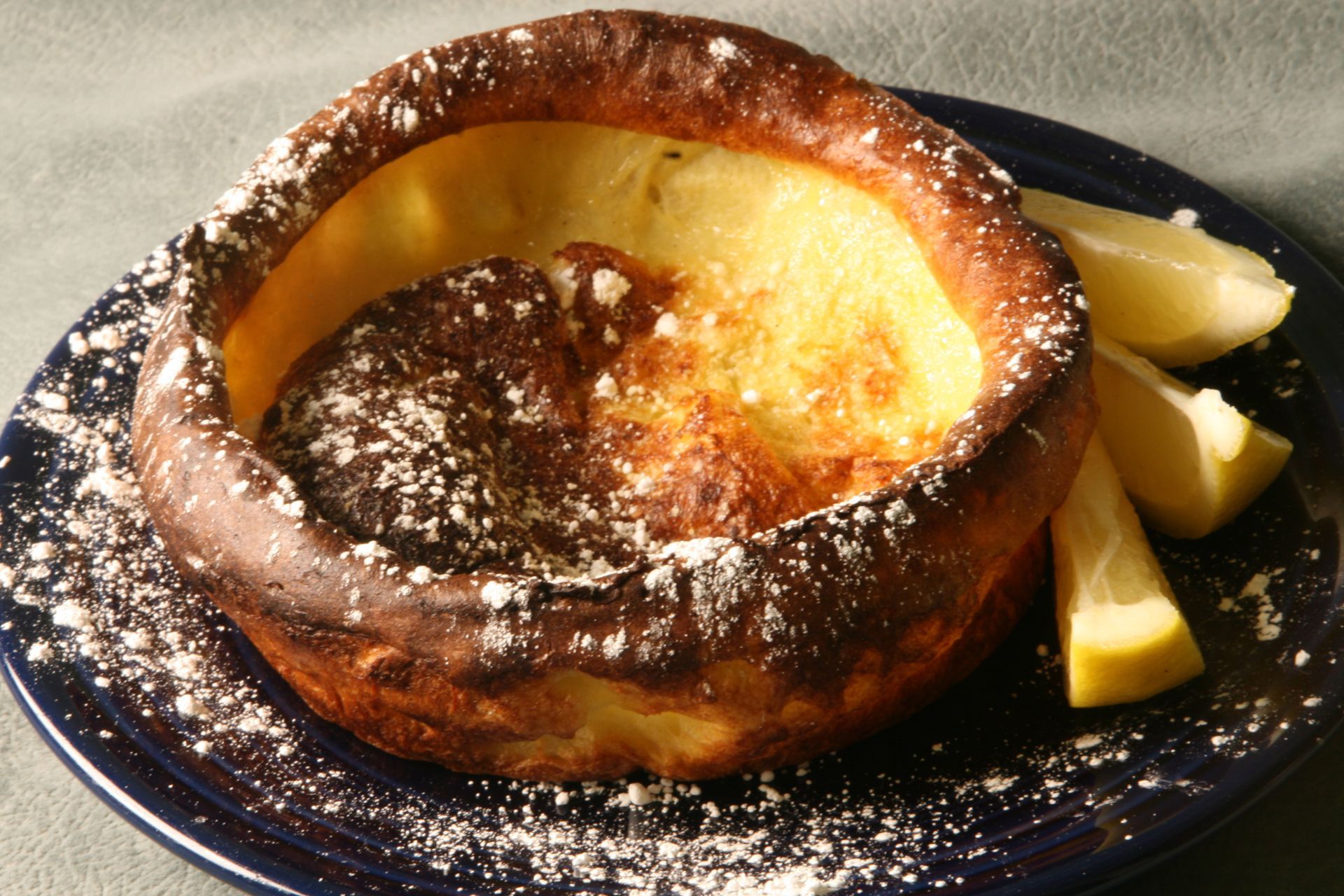 Dutch baby pancake, dusted with powdered sugar, served on blue plate with lemon wedges.
