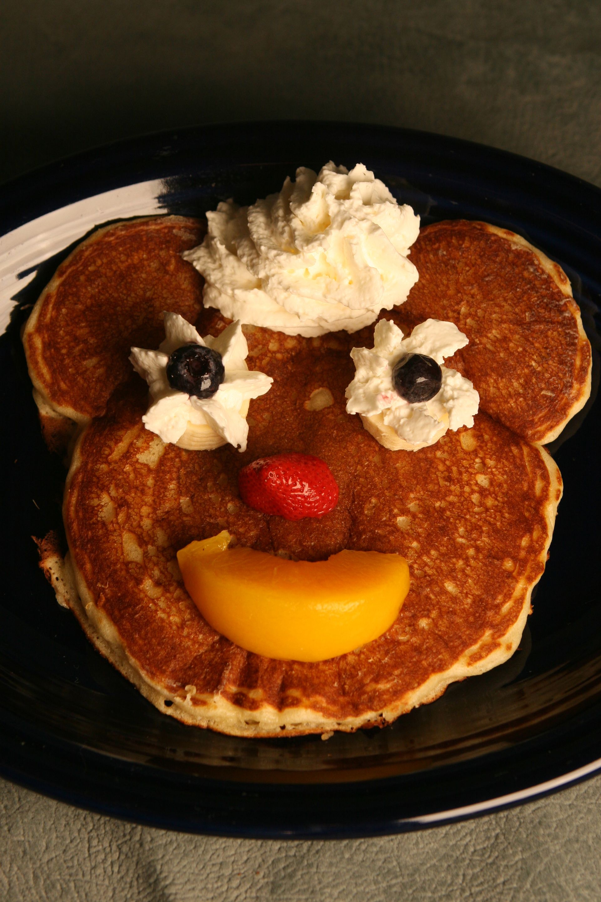 Pancakes arranged in a smiling face, decorated with whipped cream, blueberries, a strawberry, and a peach.