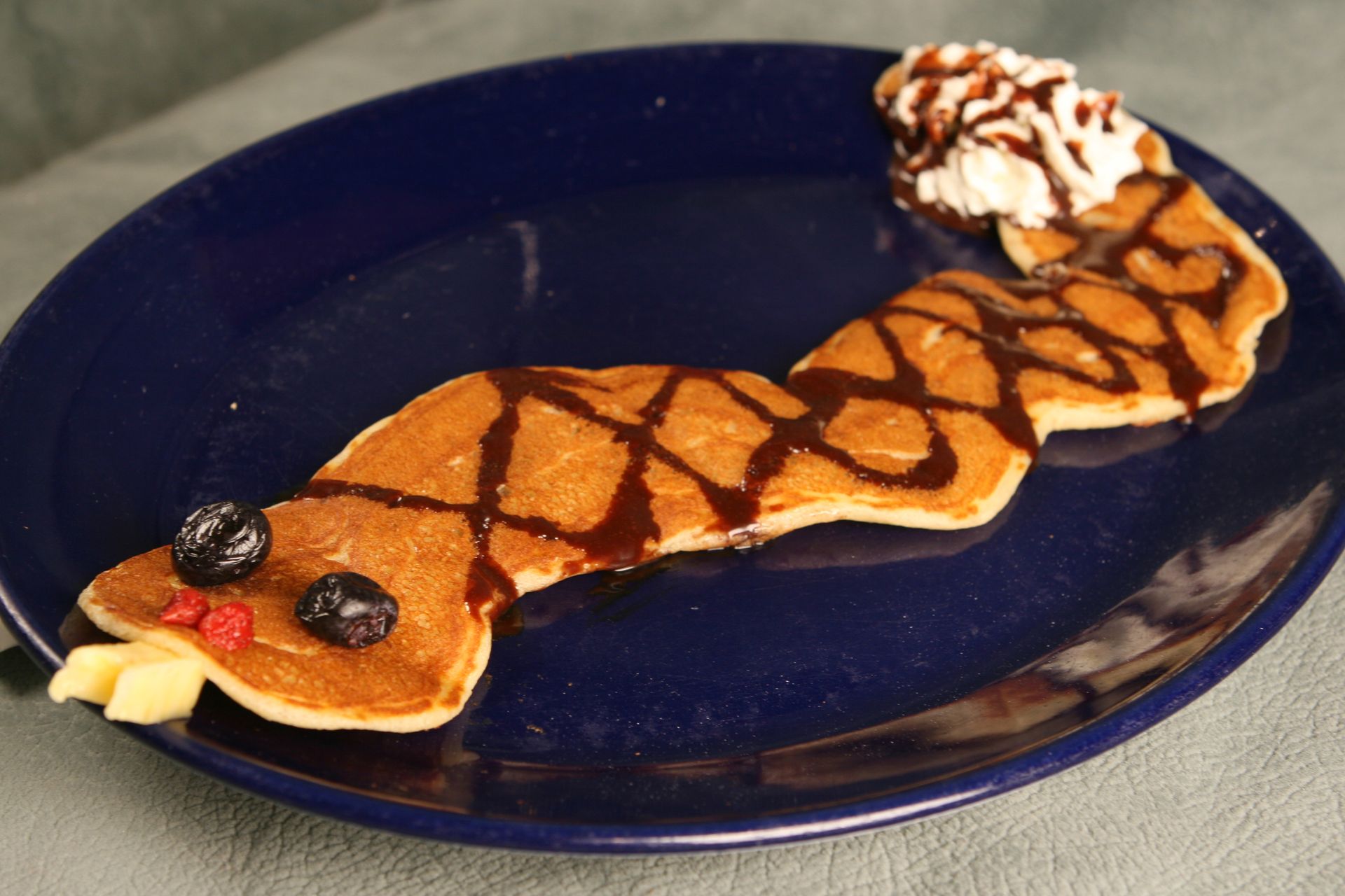 Pancake shaped like a snake on a blue plate, decorated with chocolate syrup, blueberries, and whipped cream.