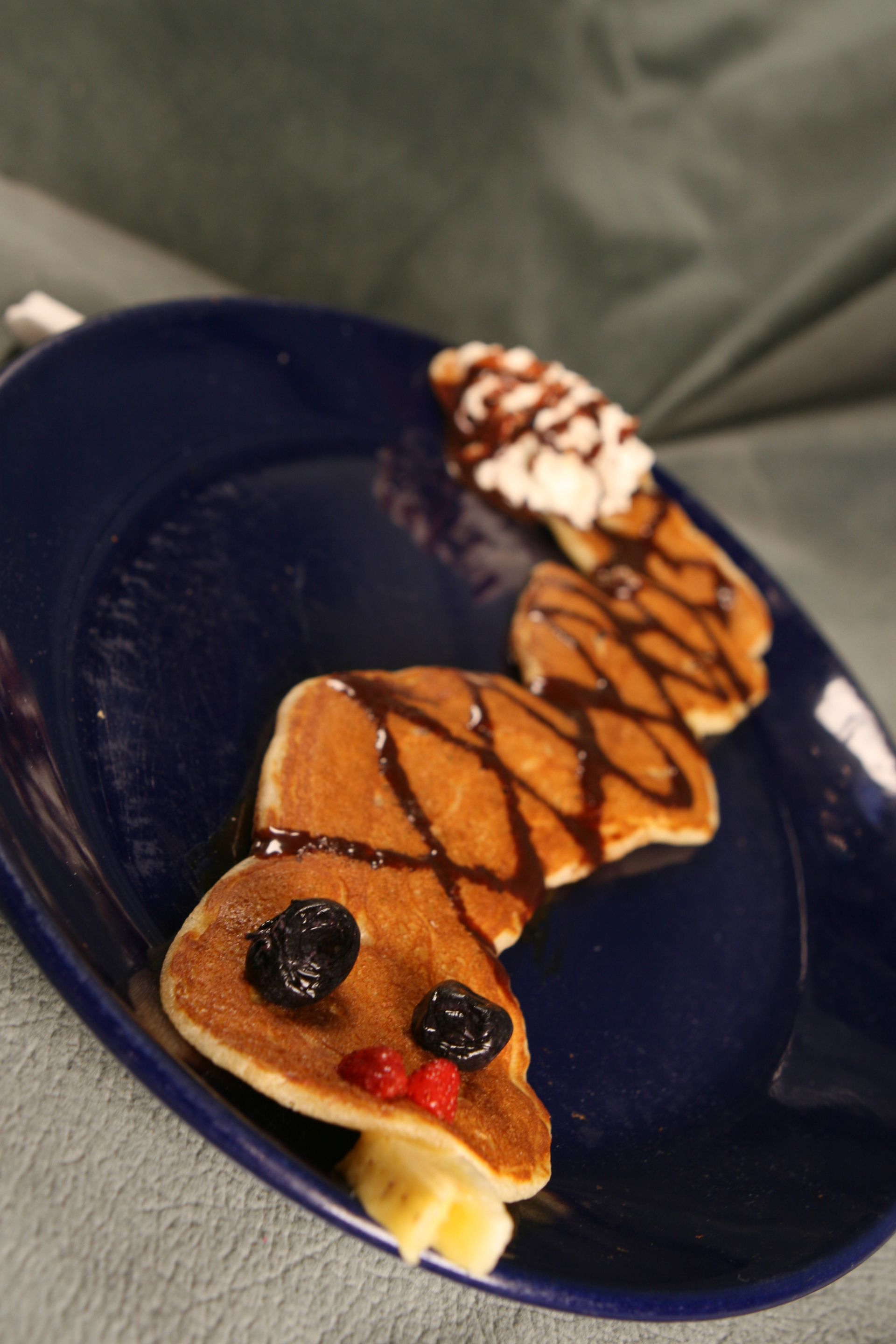 Pancake snake on a blue plate, decorated with syrup, whipped cream, and fruit.