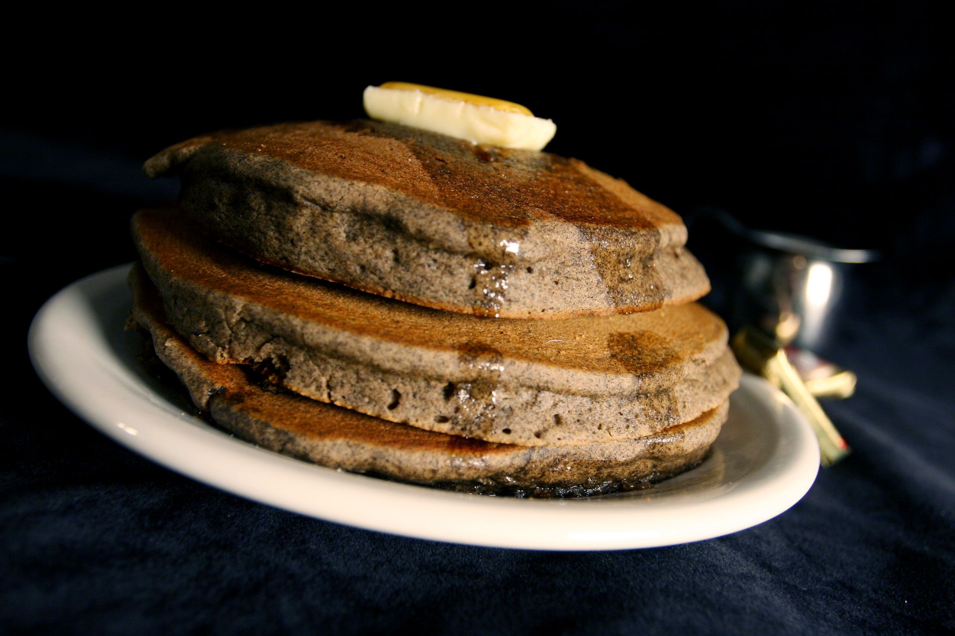 Stack of gray pancakes, butter, syrup on white plate, dark background.