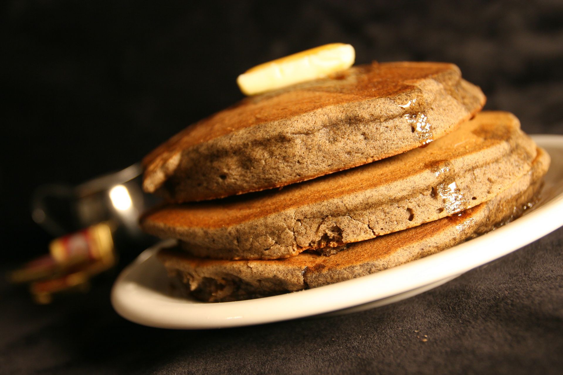 Stack of brown pancakes on a white plate, drizzled with syrup and a pat of butter, with a syrup pitcher visible.