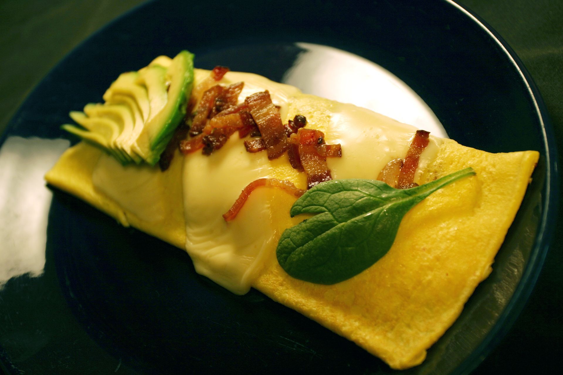 Yellow omelet topped with cheese, bacon bits, and avocado slices on a blue plate.