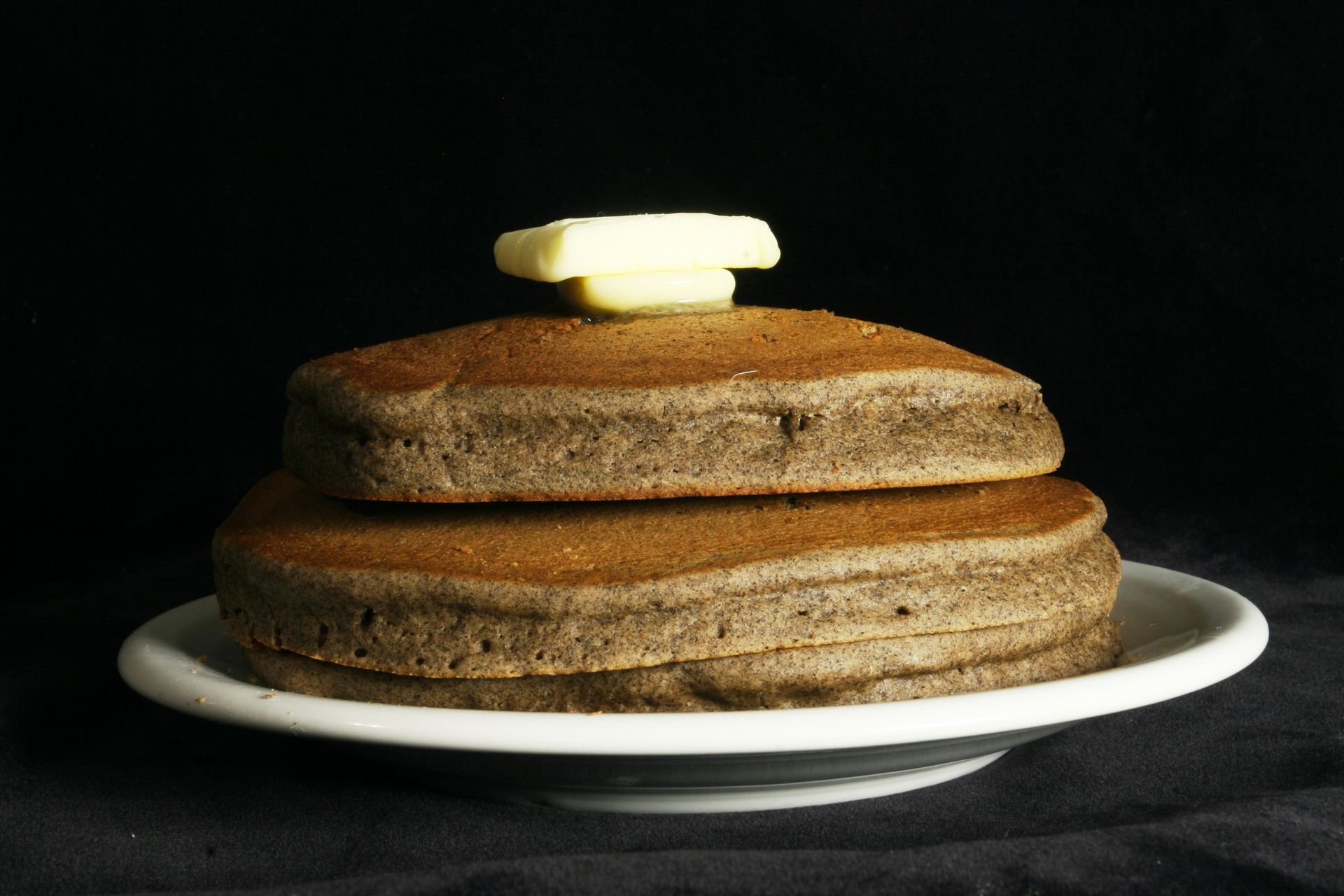 Stack of brown pancakes on white plate, topped with butter. Black background.