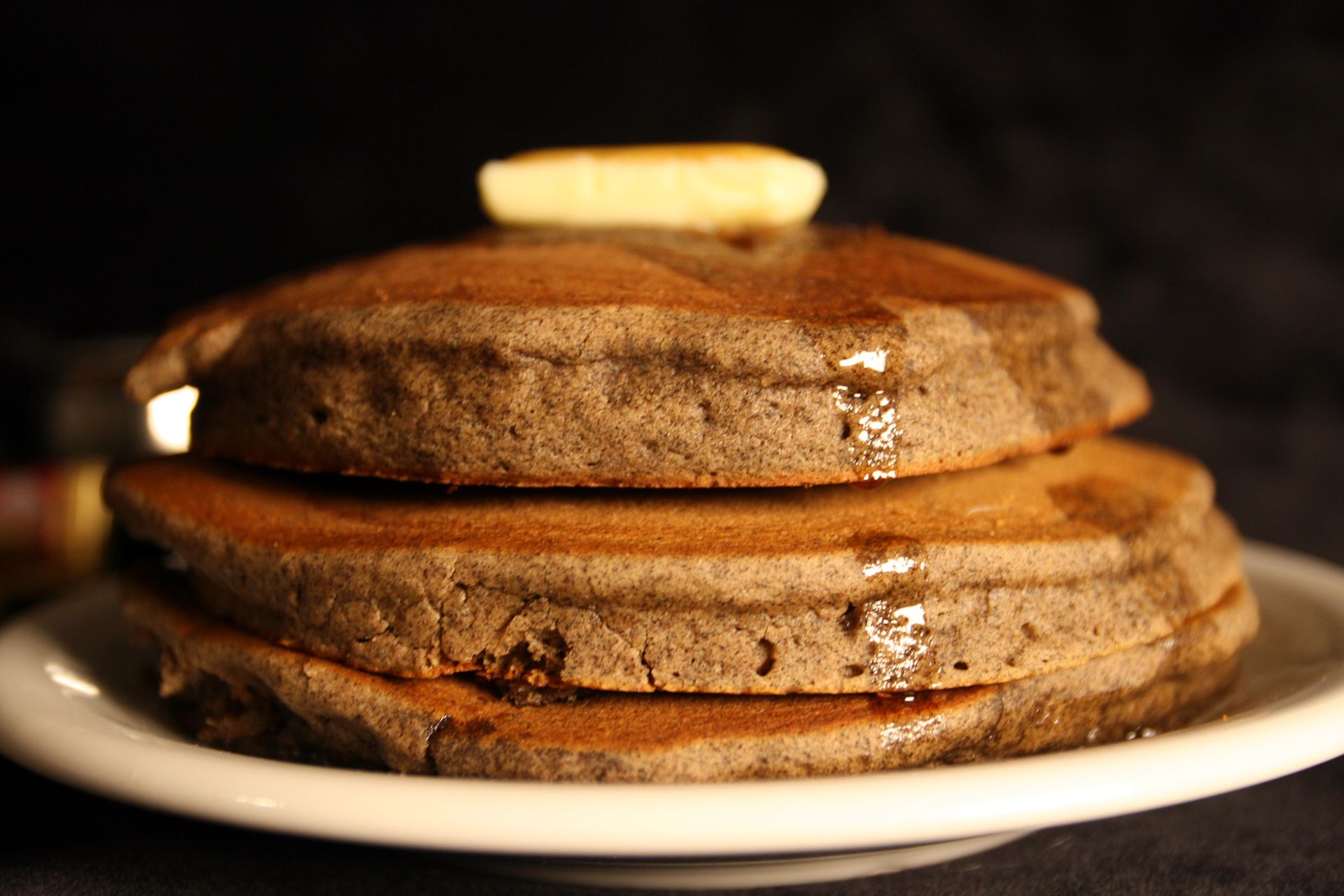 Stack of three dark pancakes on a white plate, topped with butter and syrup.