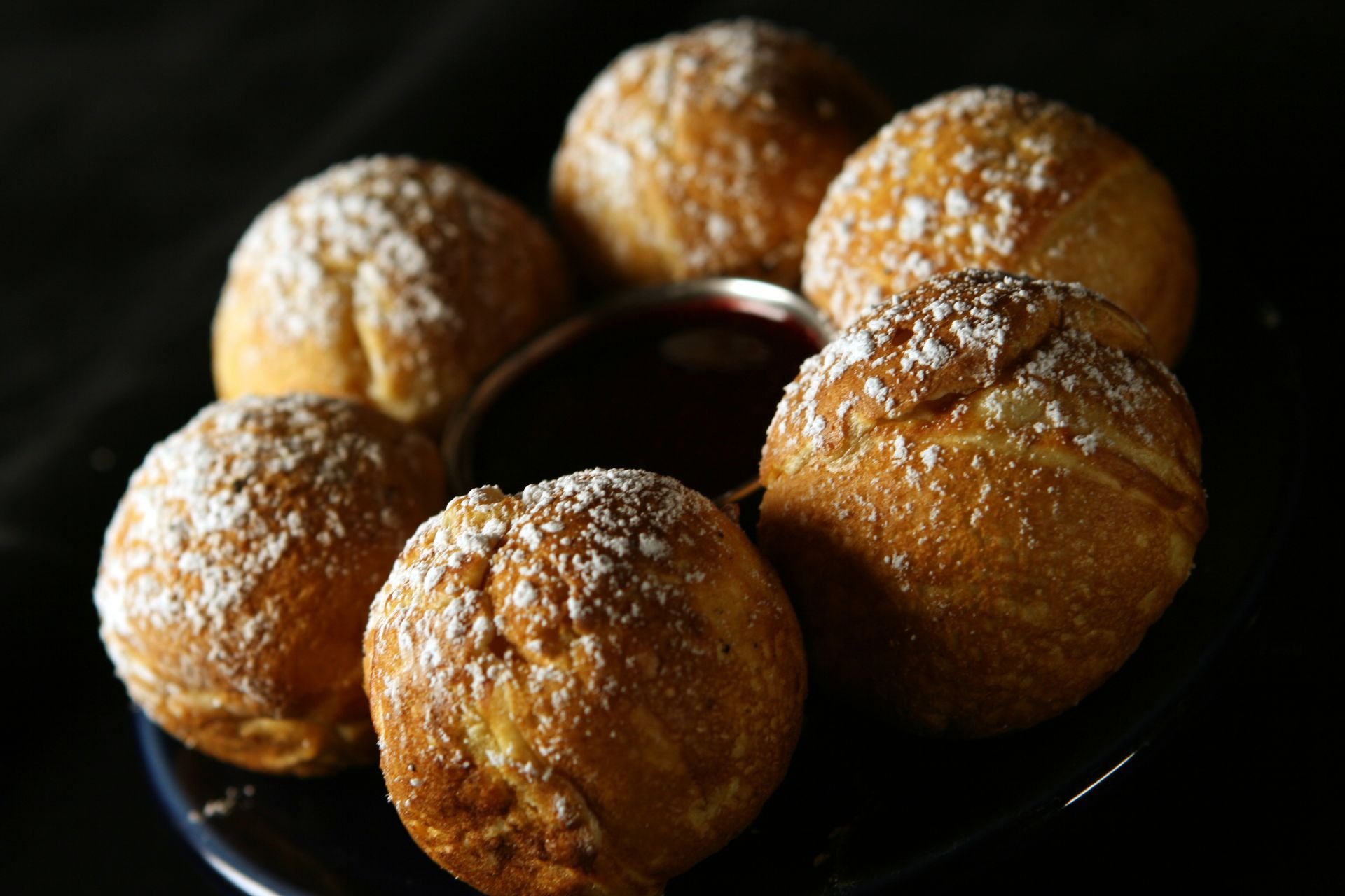 Six golden beignets dusted with powdered sugar, arranged around a dark dipping sauce on a black plate.