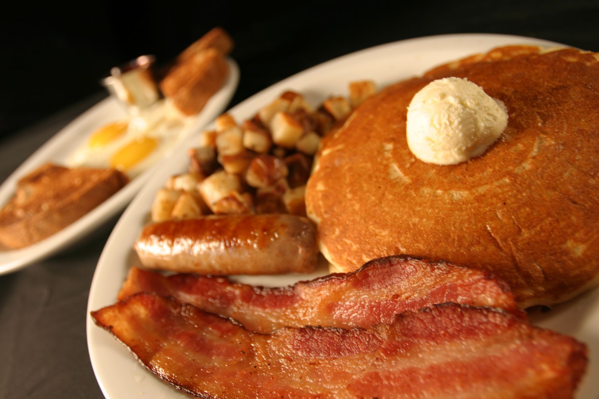 Breakfast plate: pancakes with butter, bacon, sausage, home fries, eggs, and toast.