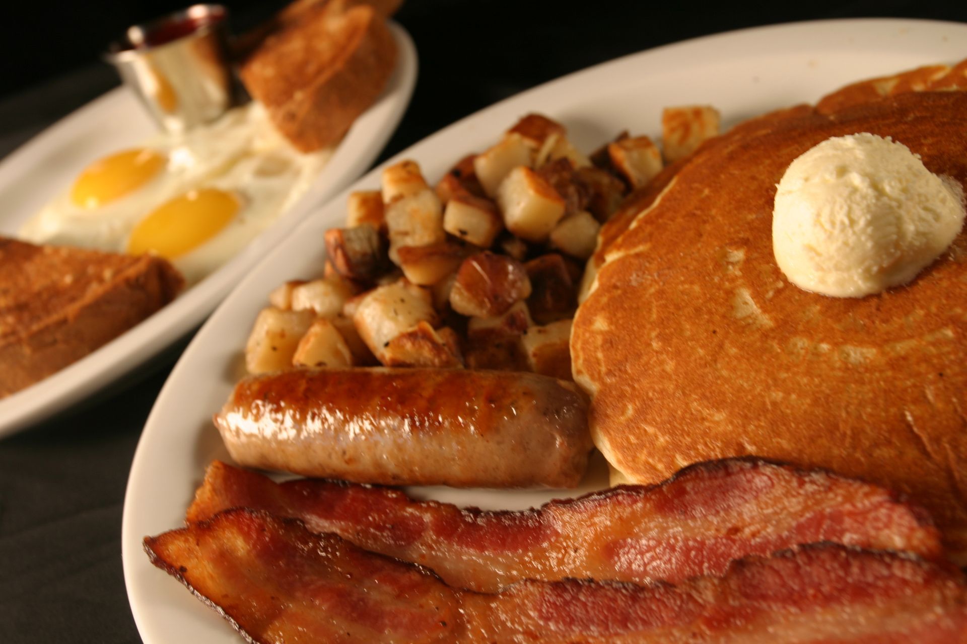 Breakfast plate with pancakes, sausage, bacon, potatoes, and butter. Eggs, toast, and syrup in the background.