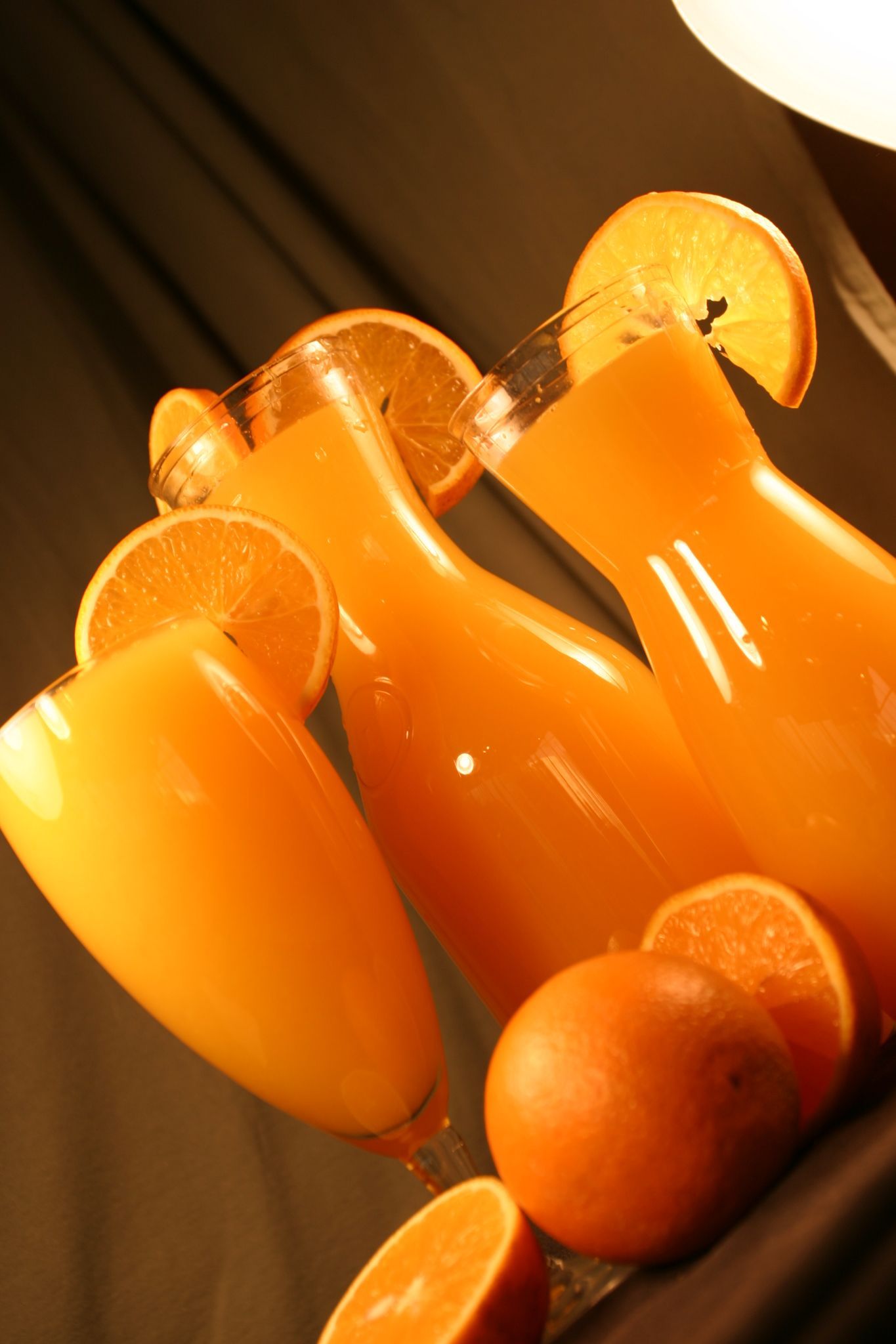 Three glasses of orange juice, garnished with orange slices, near whole and sliced oranges.