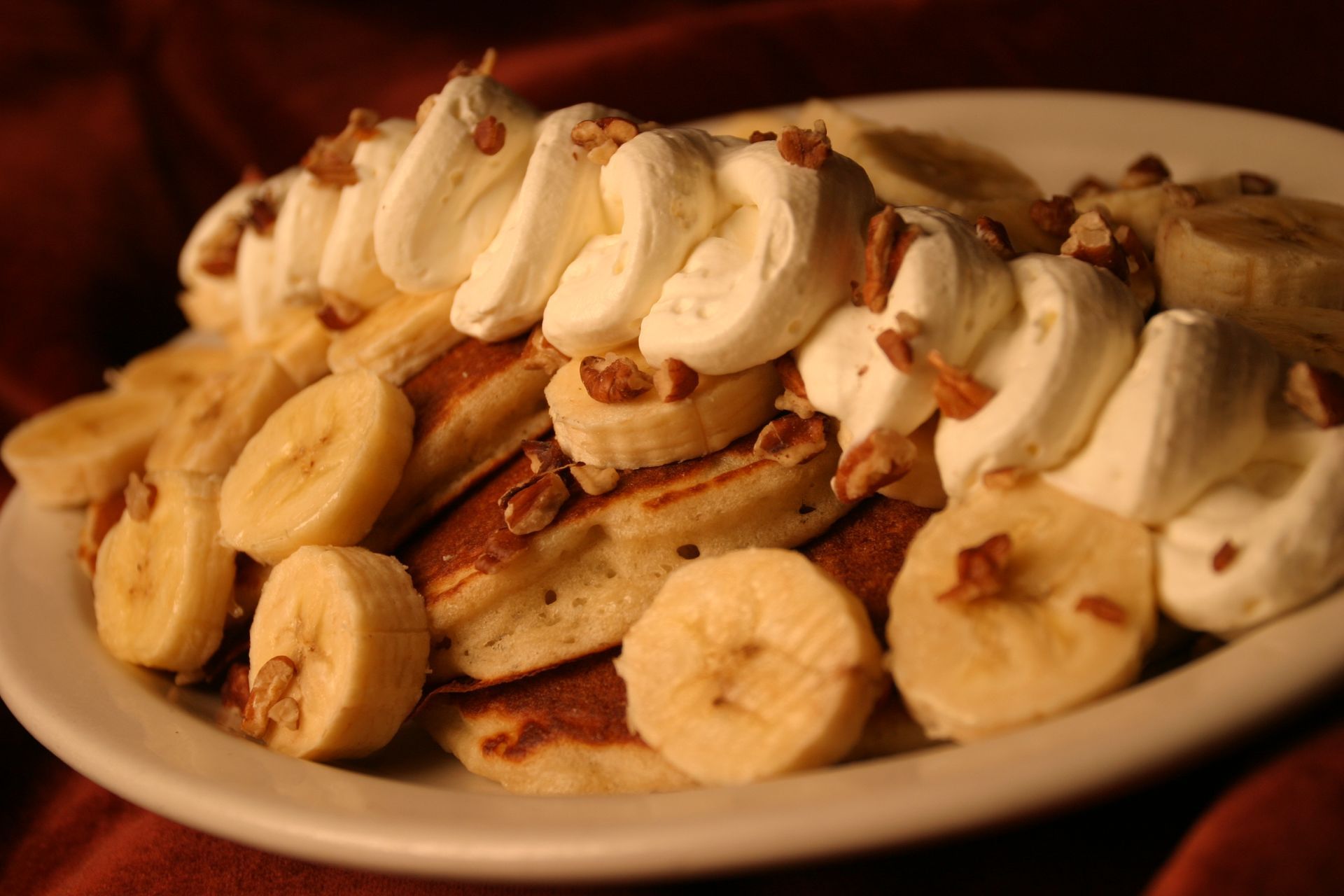 Pancakes topped with banana slices, whipped cream, and chopped pecans, on a white plate.