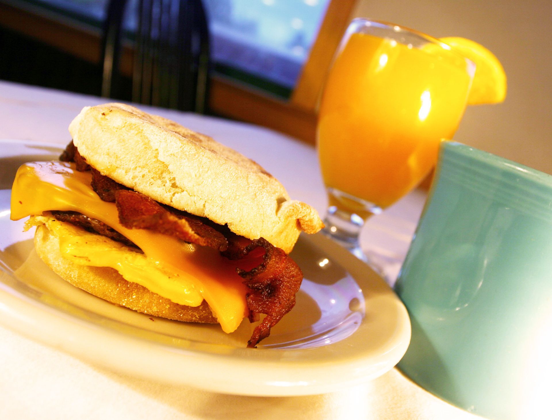 Breakfast sandwich with bacon, egg, and cheese, served with orange juice and a blue mug.