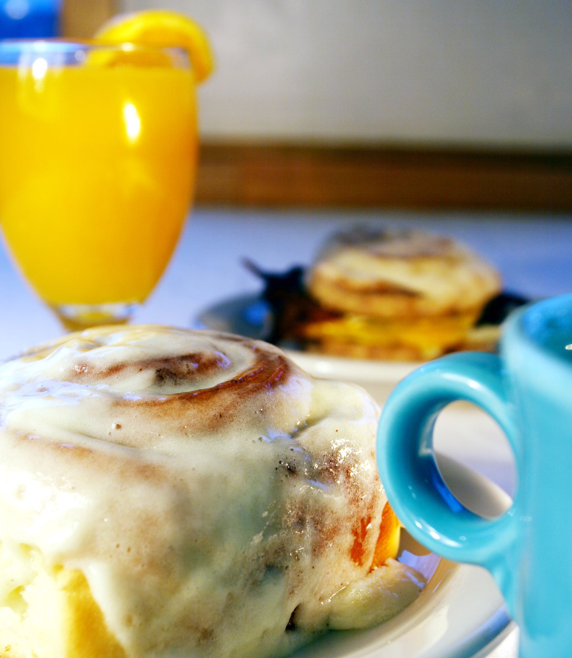 Cinnamon roll with white icing, orange juice, and a breakfast sandwich on a white plate with a blue mug.