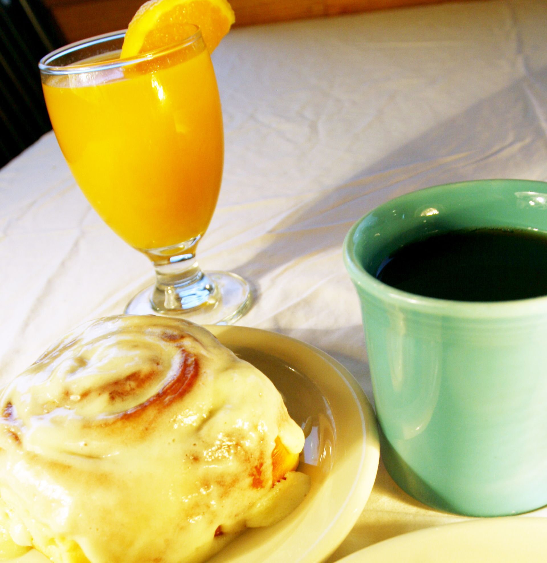 Cinnamon roll, orange juice with garnish, and coffee in a turquoise mug on a white tablecloth.