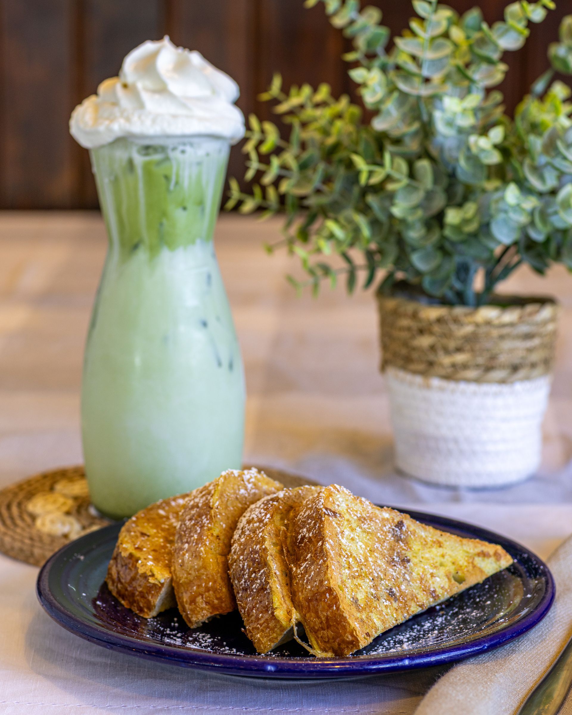 French toast on a plate and matcha drink with whipped cream, near potted plant.