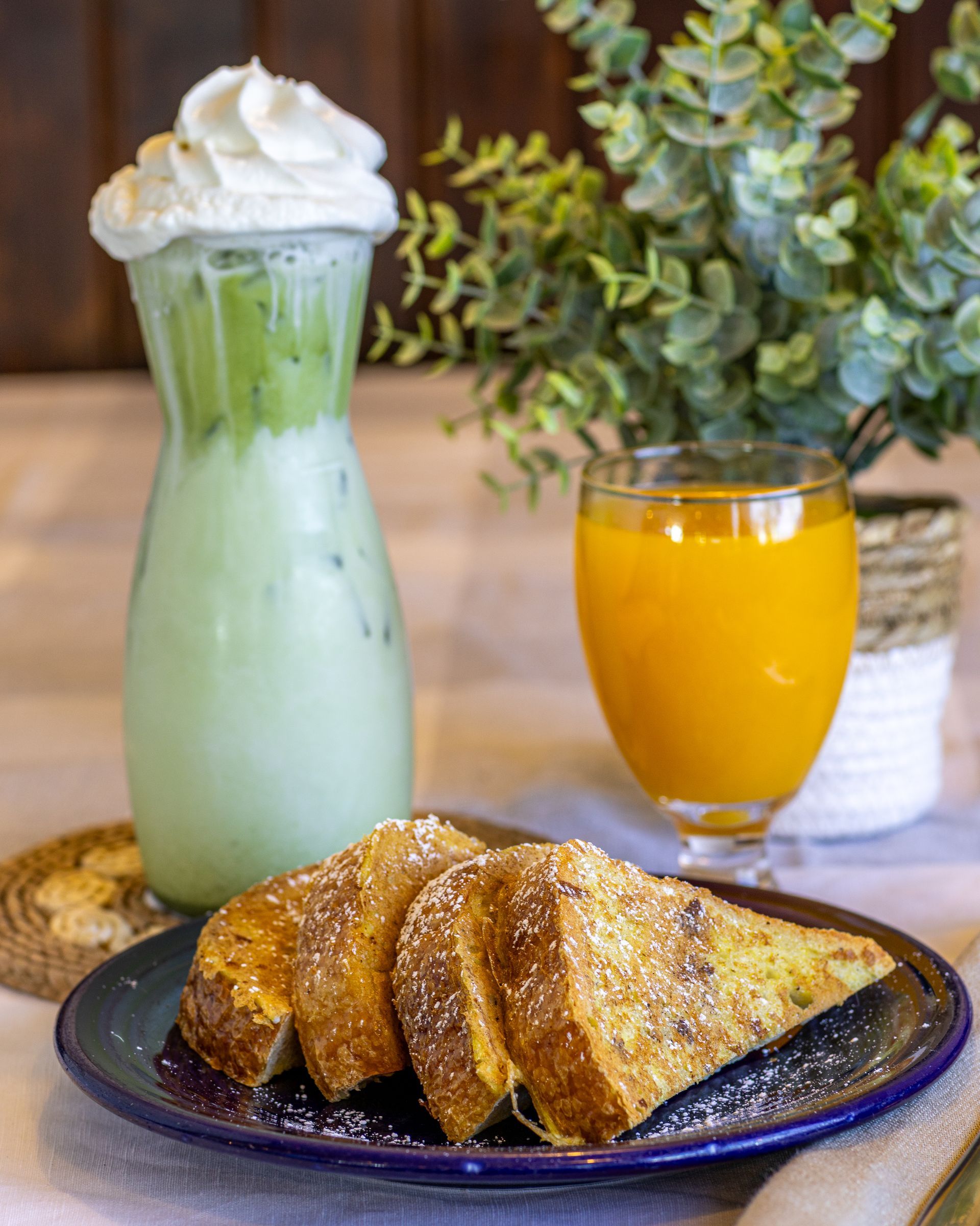 French toast slices on a plate with matcha drink, orange juice, and a plant in the background.