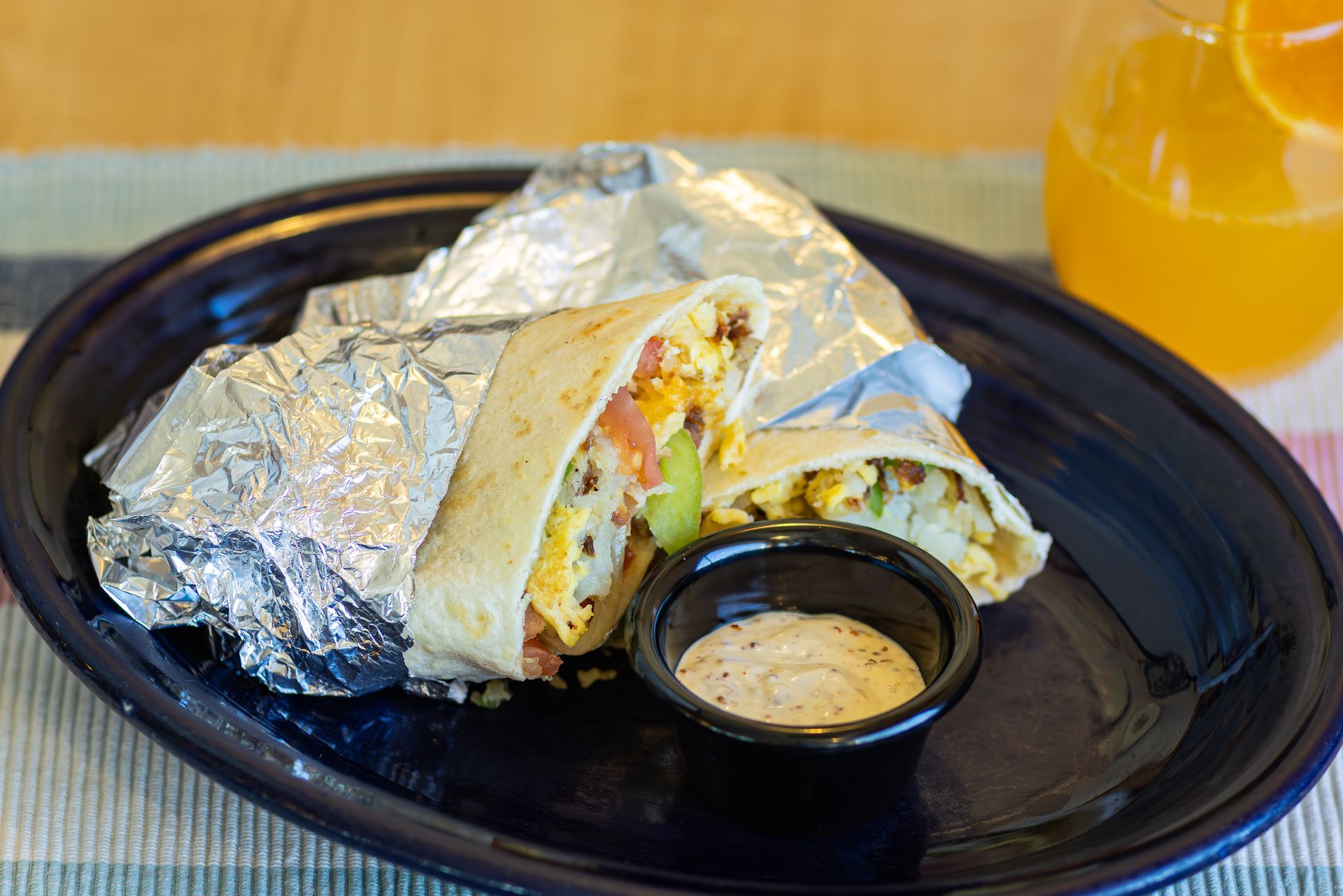 Breakfast burrito, cut in half, with dipping sauce on a plate, alongside a glass of orange juice.