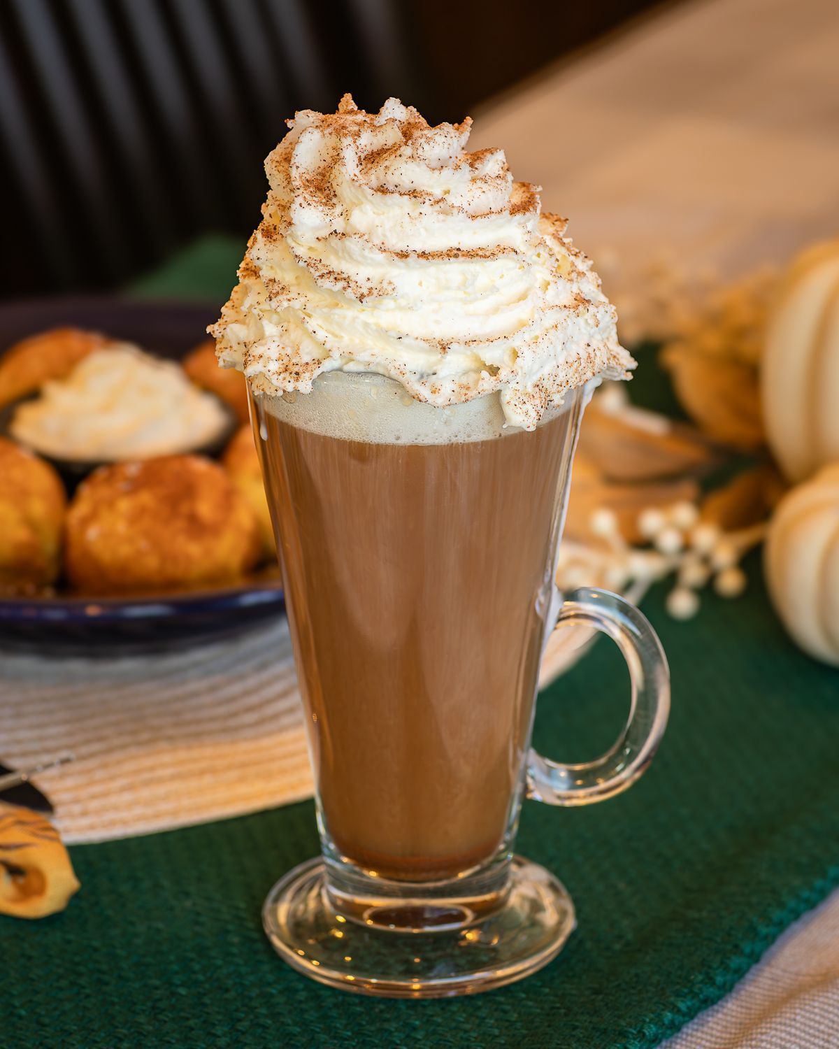 Tall glass of hot chocolate topped with whipped cream and cinnamon. Beside plate of golden puffs, pumpkins in the background.