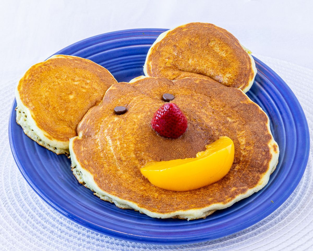 Bear-shaped pancakes with a strawberry nose, peach smile, and chocolate chip eyes on a blue plate.