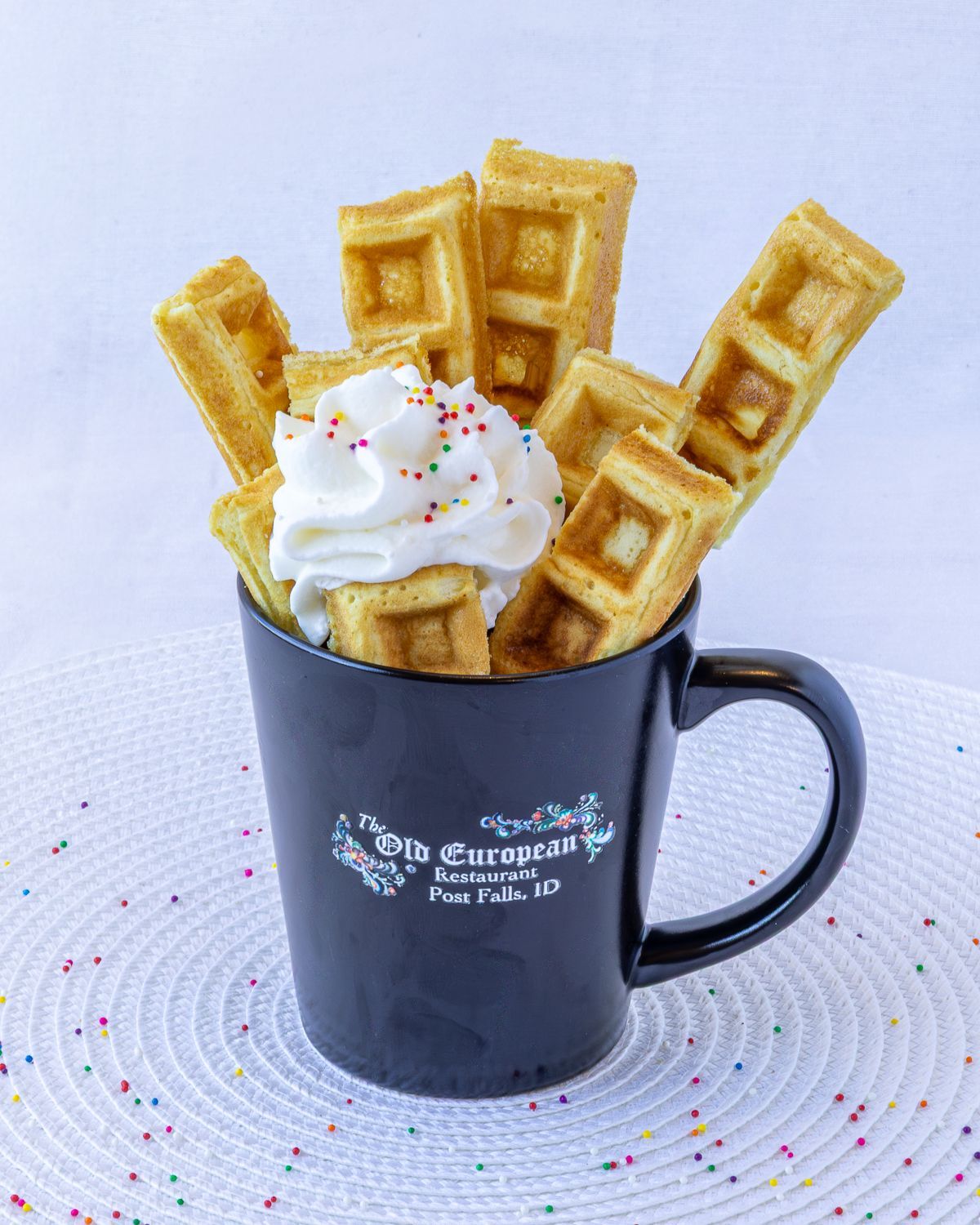 Waffle sticks in a black mug with whipped cream and sprinkles, on a white textured mat.