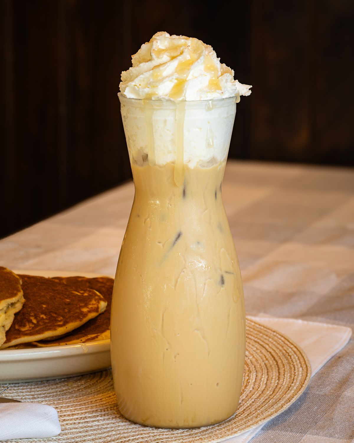 Iced coffee in a decorative glass, topped with whipped cream and drizzle. Pancakes on the side.