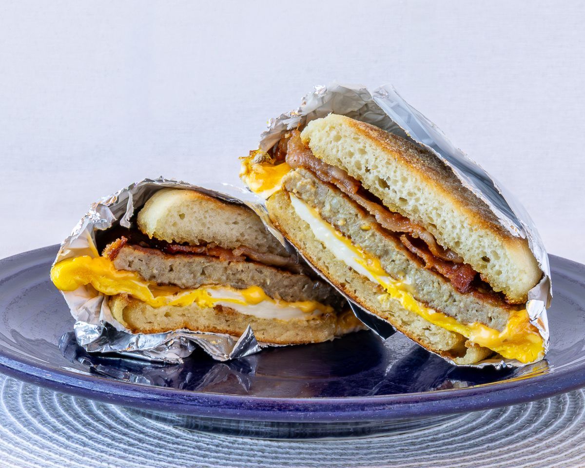 Breakfast sandwich halves wrapped in foil, on a blue plate. Includes egg, bacon, sausage, and bread.
