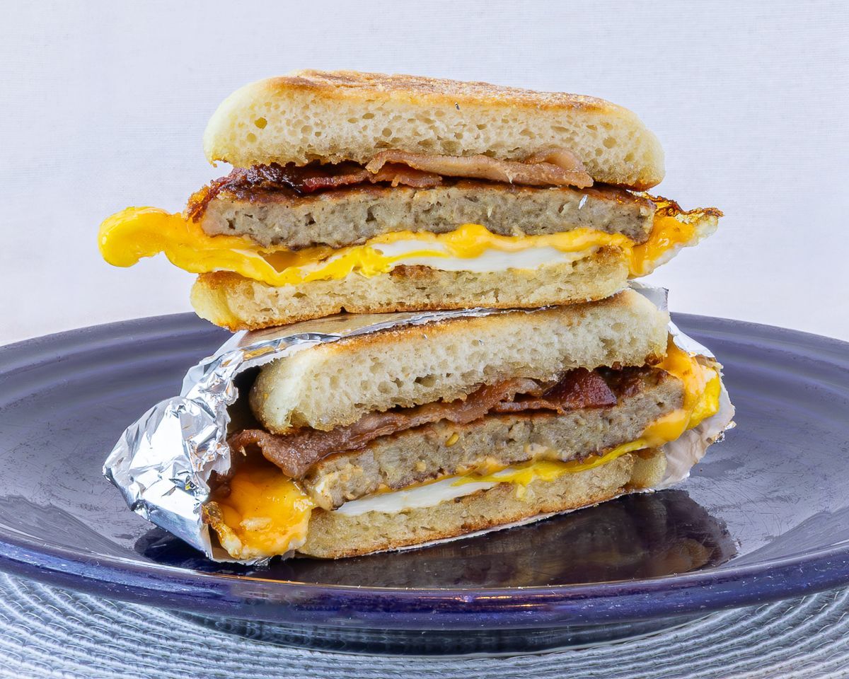 Breakfast sandwich stacked with egg, sausage, bacon, on toasted bread, wrapped in foil, on a blue plate.