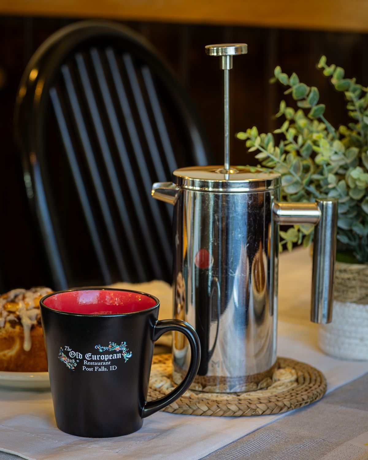 Coffee press and mug on a table; behind, a chair and plant. Mug is black with red interior.