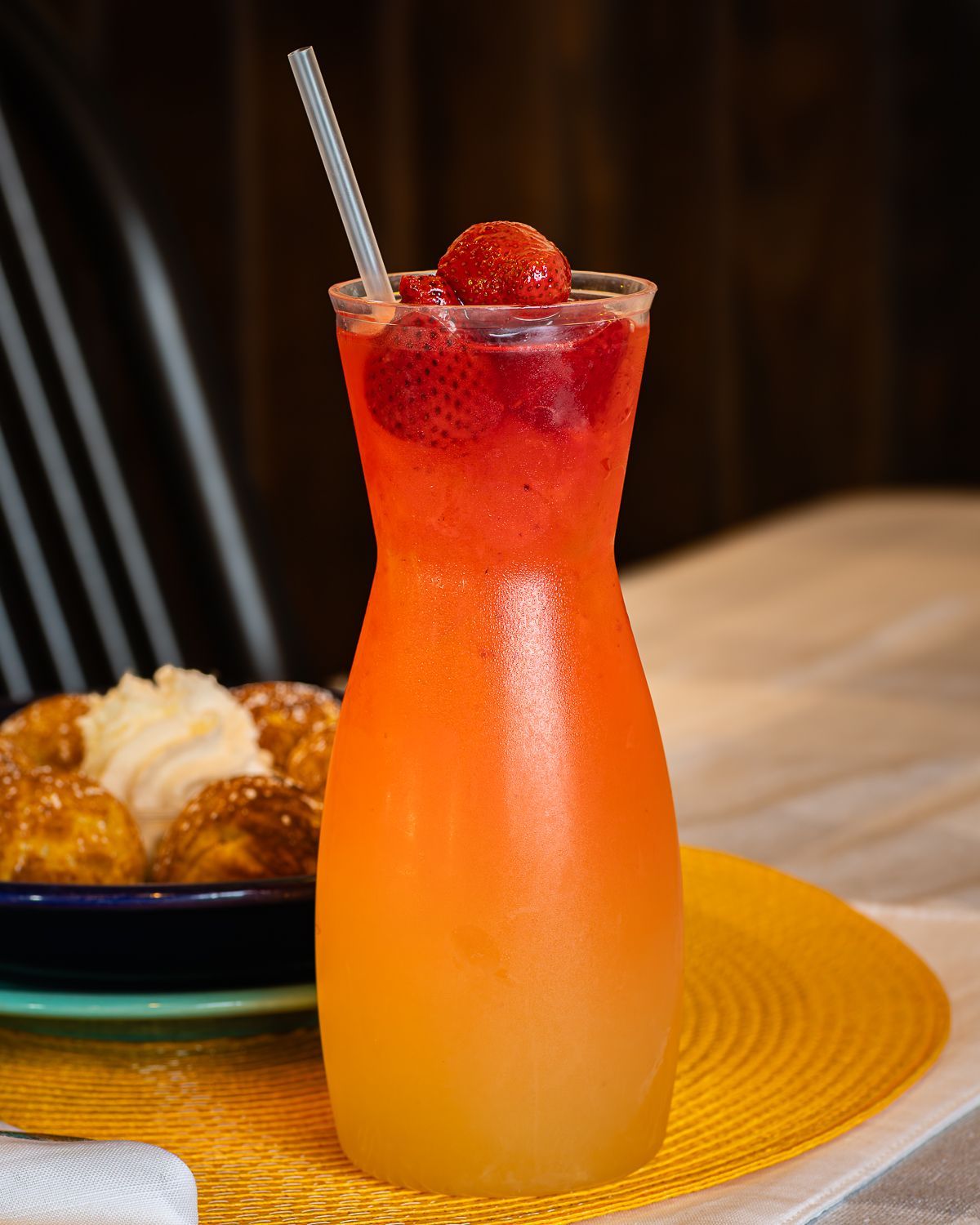 Sunrise-colored drink in a tall glass with a straw, garnished with strawberry, on a yellow mat next to pastry.