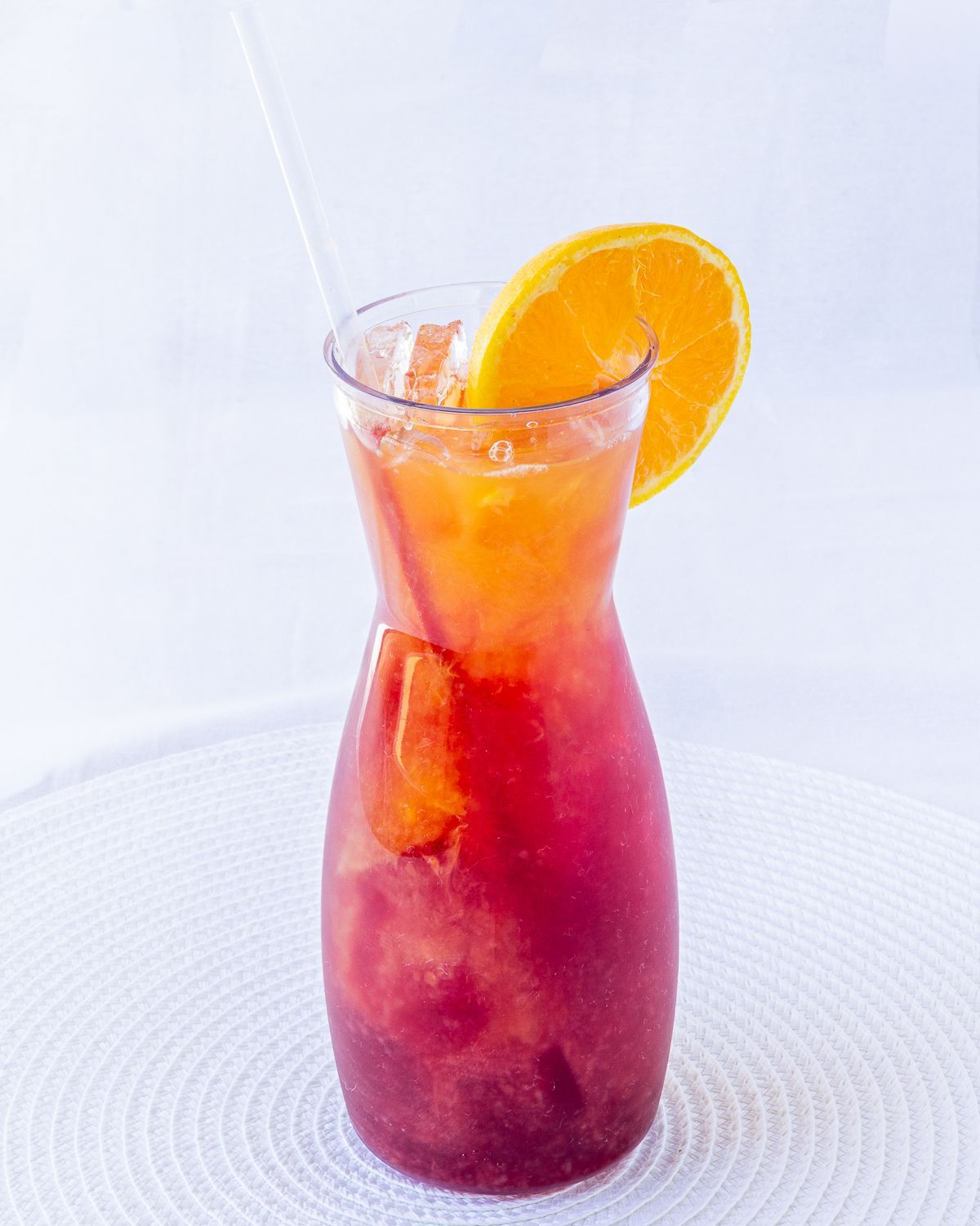 A tall, layered cocktail in a glass with an orange slice and straw on a white surface.