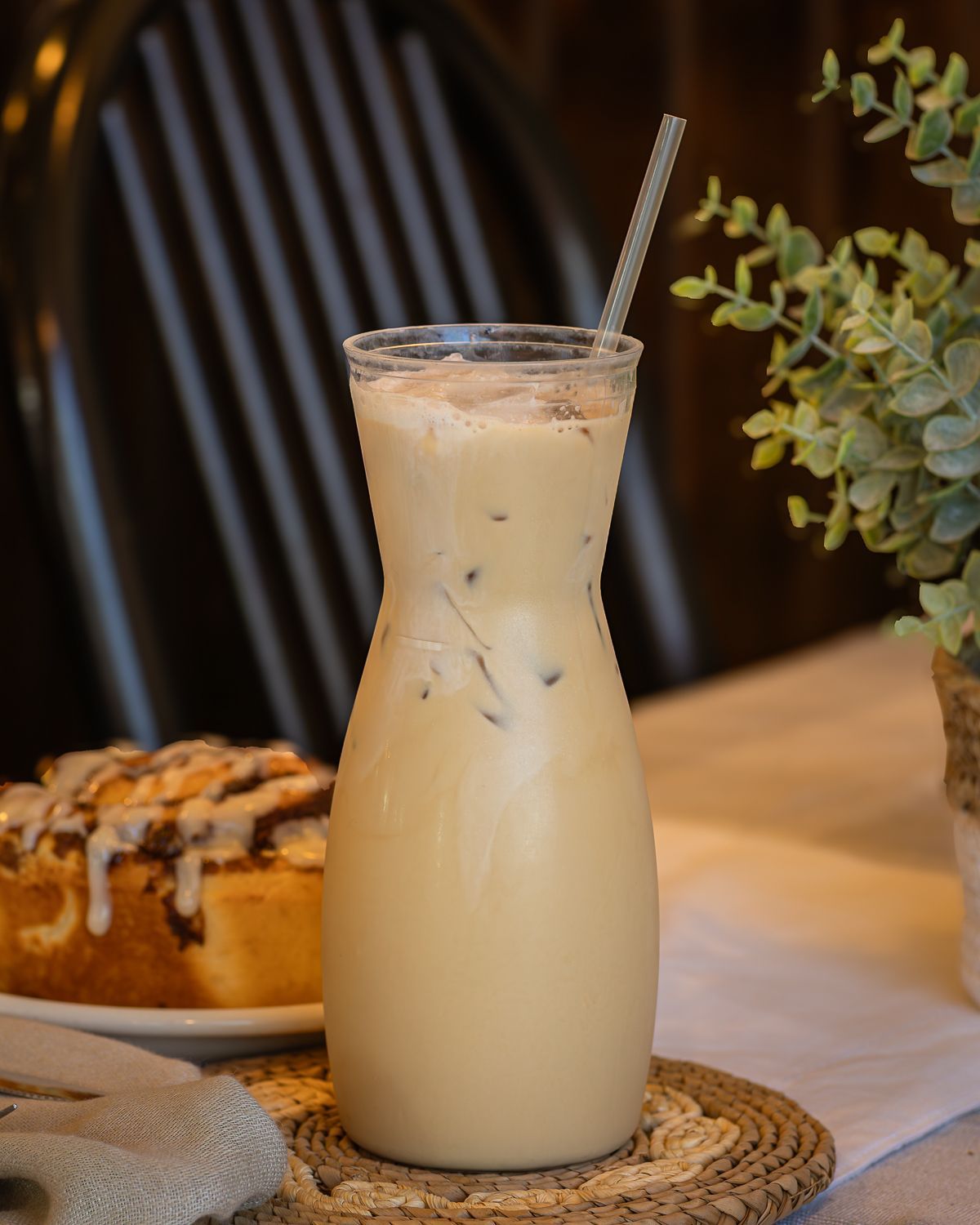 Iced coffee in a tall glass with straw, beside a cinnamon roll and plant.