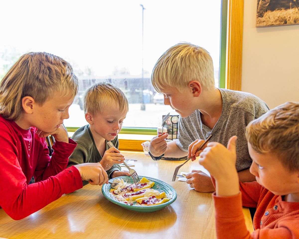 Four children eating at a table by a window, focused on food on a shared plate.